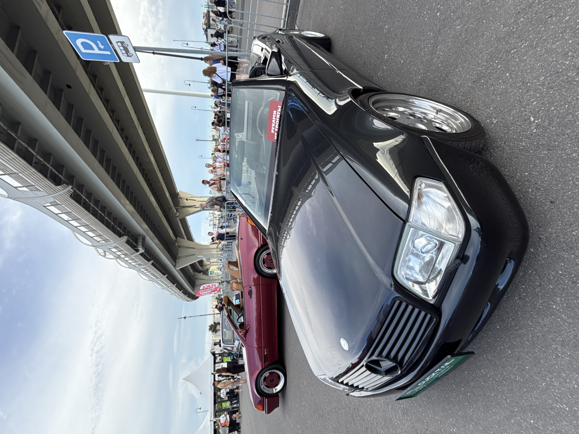 Two Mercedes-Benz convertible cars are displayed outdoors. In the foreground is a black Mercedes-Benz SL-Class (R129) with its top down. A red sign on the windshield reads "РУКАМИ НЕ ТРОГАТЬ!" (Do not touch with hands!). A green license plate frame is attached to the front bumper. Behind it, a burgundy Mercedes-Benz SL-Class (R107) is also parked with its top down. The cars are parked on asphalt, likely at an outdoor event or car show. In the background, a pedestrian overpass or elevated highway structure dominates the scene. People are gathered and standing near a metal fence to the right of the cars, looking at the vehicles. Tents are visible in the far left background. A blue and white parking sign with a P and an arrow pointing left is mounted on a pole to the right of the overpass. The sky is partly cloudy. The event appears to be taking place under an elevated transportation structure typical of urban environments, possibly in Saint Petersburg, Russia, given the architectural style of the overpass and the presence of the Russian text on the sign.