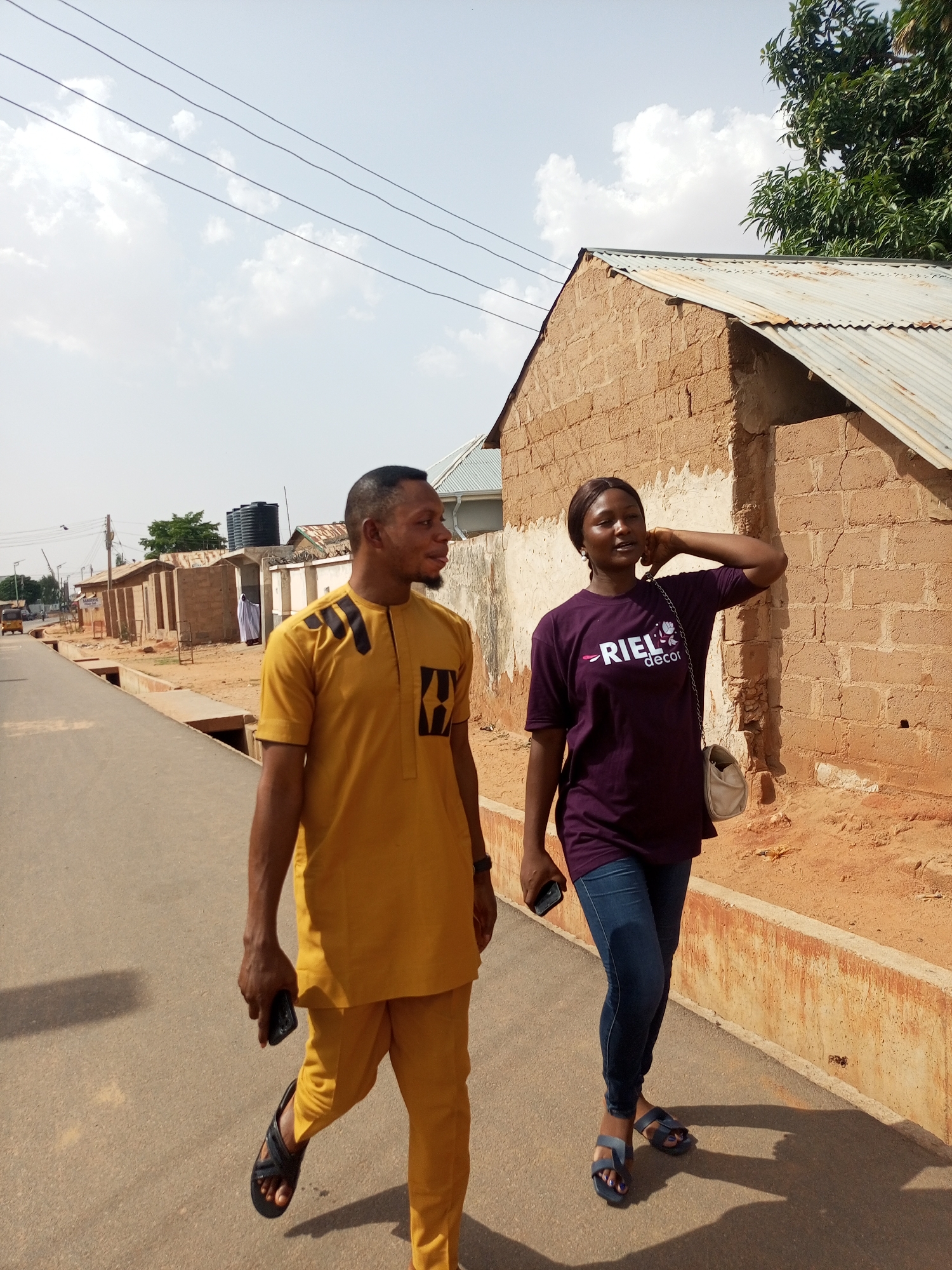Two individuals, a man and a woman, are walking on a paved street in Tempure, Nigeria. The man is on the left, wearing a yellow traditional outfit and sandals, holding a mobile phone. The woman, on the right, is wearing a purple t-shirt, jeans, and sandals, with a small purse over her shoulder, also holding a mobile phone and touching her neck. They are walking in the same direction, away from the camera. To the right, there's a low building constructed of mud bricks with a corrugated metal roof, while similar structures line the street in the background. The sky is partially cloudy. Power lines stretch across the sky. There is a small amount of green vegetation in the background. A vehicle is driving down the street in the distance.