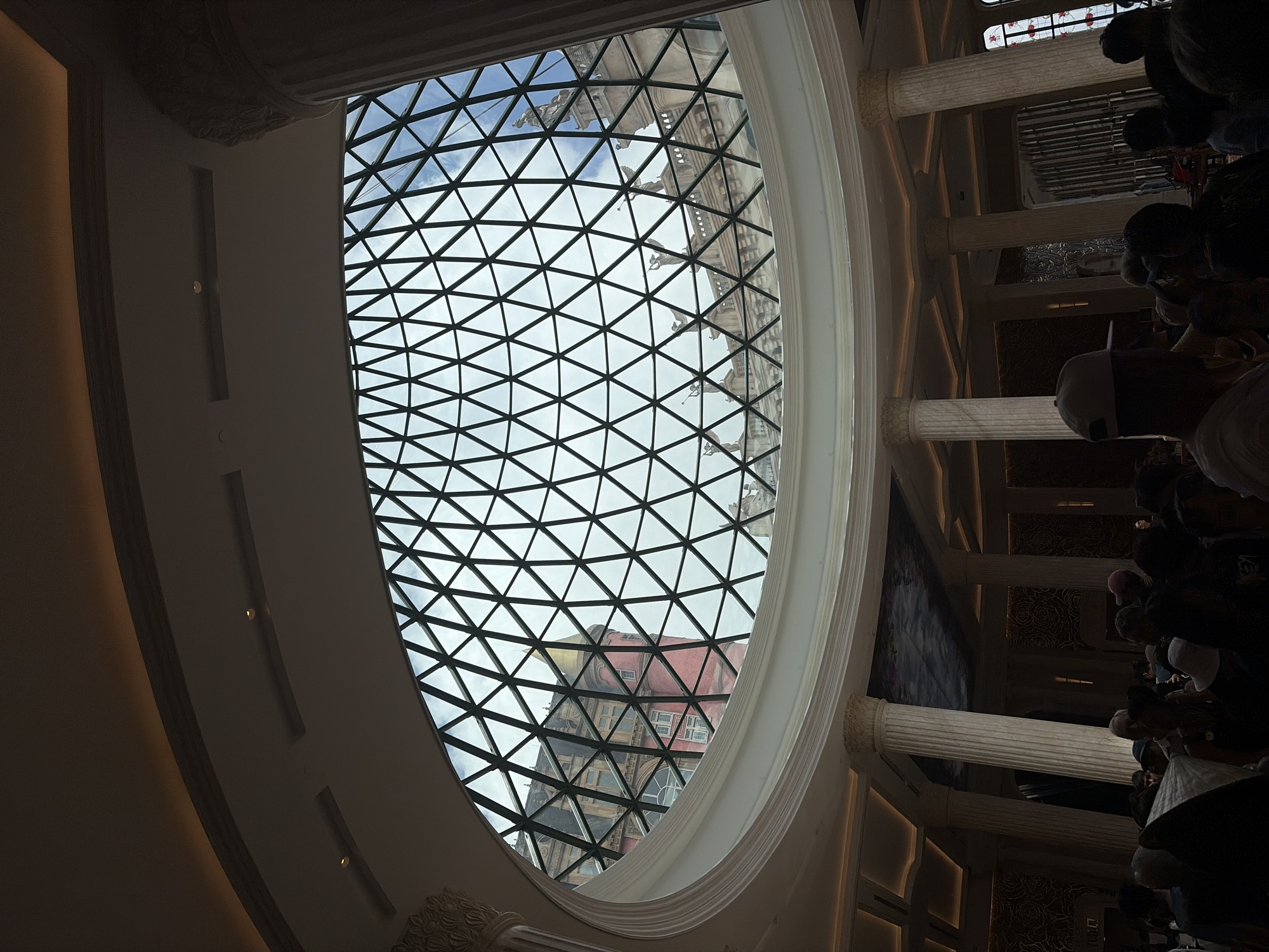 The image shows a view looking up through a glass dome with a triangular grid pattern. The dome is part of a building with classical architectural elements, including columns and ornate moldings. Through the dome, another building with a red and tan facade is visible. On the right side of the image, there are columns and people. The image is taken from inside a building with classical architecture. The location appears to be Đà Nẵng, Vietnam, based on the architectural style.