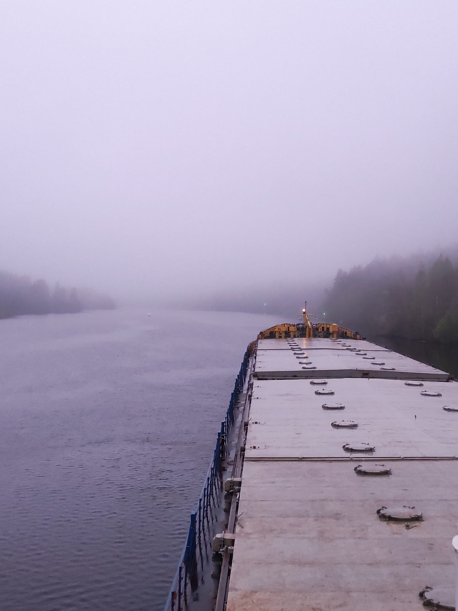 A forward-facing, eye-level perspective of a large barge transiting a waterway, heavily obscured by dense fog. The barge occupies the right half of the frame, its deck composed of rectangular segments with evenly spaced circular hatches. At the rear of the barge, a yellow structure with antennae-like protrusions is visible. The waterway occupies the left half of the frame, its surface reflecting the diffused light of the foggy atmosphere. The far bank of the waterway is faintly visible through the fog, marked by the silhouette of trees. The fog obscures the horizon. This is likely Volokov Most, Russia, based on the waterways in the area.