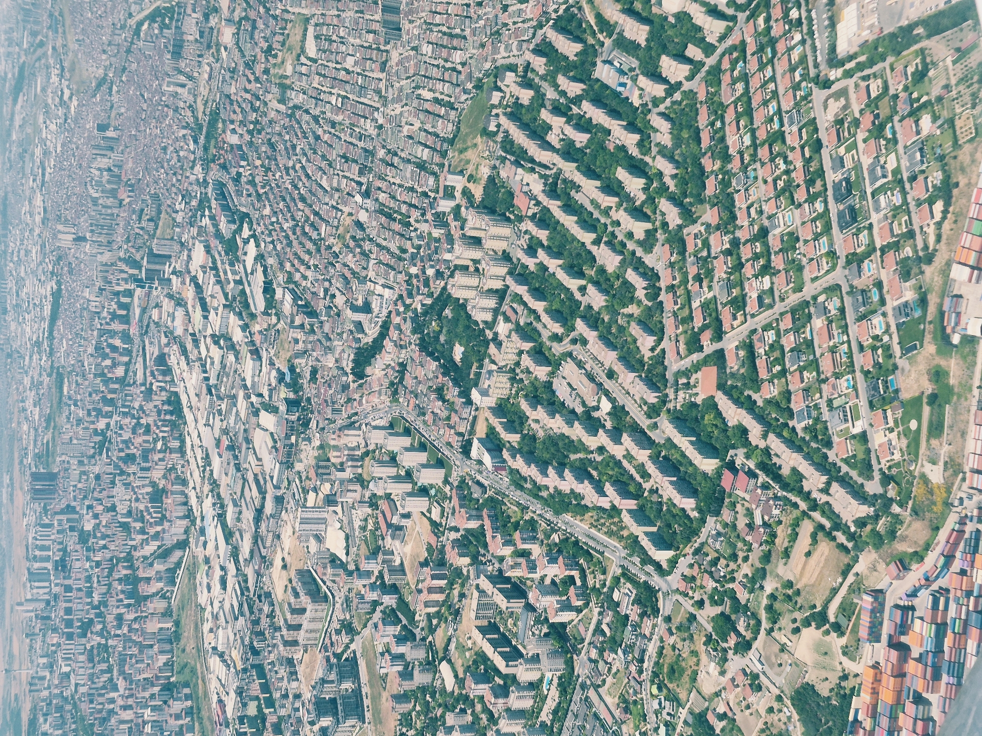 This is an aerial photograph of a dense urban landscape, identified as Beylikdüzü, Turkey. The image is dominated by an extensive grid of residential buildings, varying in size and architectural style. In the foreground, a collection of stacked shipping containers is visible, suggesting proximity to a port or industrial area. The midground features a uniform development of mid-rise apartment buildings, many with red-tiled roofs, arranged in rows with tree-lined streets and courtyards. Further into the background, the cityscape transitions to a mix of taller residential towers and commercial or industrial buildings, with a sprawling network of roads connecting them. The overall impression is one of high-density urban living with pockets of green spaces interspersed among the structures. There are no visible individuals or specific actions depicted in the photograph; it is a static overview of an urban environment. The lighting suggests daytime, possibly late morning or early afternoon, given the shadows cast by the buildings. The sky is hazy, which is common in urban areas.
