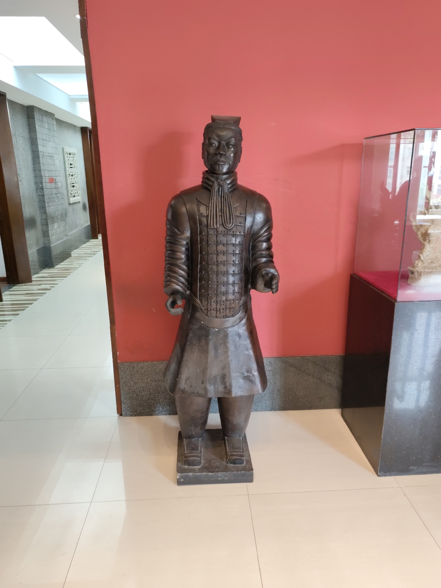A life-sized, dark-colored bronze statue of a Terracotta Warrior stands in the foreground, facing forward. The warrior is depicted wearing armor with intricate detailing on the chest and shoulders, a pleated skirt, and a distinctive headdress. The statue is positioned on a dark rectangular base. Behind the statue, a vibrant red wall serves as the backdrop. To the right of the statue, a glass display case is partially visible, with a red velvet cloth lining the bottom and what appears to be a light-colored, textured object inside. To the left of the statue, a hallway extends into the distance, featuring a grey tiled wall on the left side with a decorative white inlay. The floor in the foreground is tiled with large, light-colored square tiles. The scene appears to be indoors, possibly in a museum or exhibition space, in 幸福乡, China.