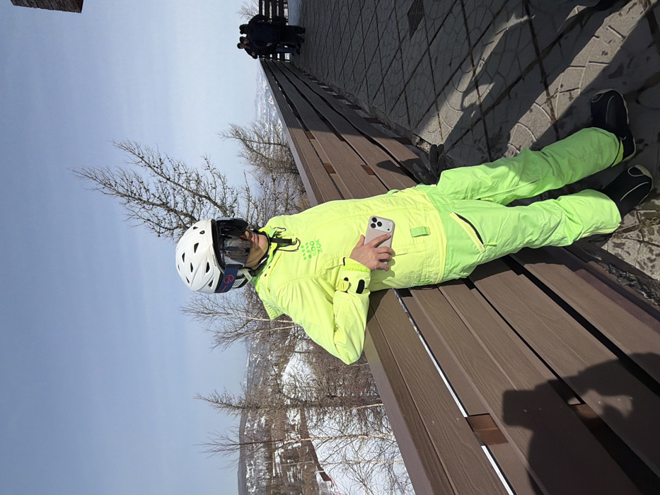 An adult female in full ski attire is depicted standing on a paved observation deck in Zelenaya Polyana, Russia, overlooking a snowy mountain landscape under a clear sky. The subject wears a white ski helmet with dark, mirrored goggles covering her eyes, a fluorescent yellow-green hooded ski jacket with black detailing and "COO ZOK" text, and matching ski pants with black accents and zippered pockets. Her lower legs are obscured by black ski boots or liners. She is leaning with her left arm resting on a dark brown wooden railing that extends along the deck. In her right hand, she holds a light-colored smartphone with a triple-camera system, positioned against her hip. In the far right background, several other individuals in winter clothing are standing on the same paved surface beyond the railing. The surrounding environment consists of snow-covered slopes, sparse deciduous trees with bare branches, and some evergreens. A marked ski run is visible in the mid-ground to the left. The ground surface is composed of grey rectangular pavers, and bright sunlight casts distinct shadows.