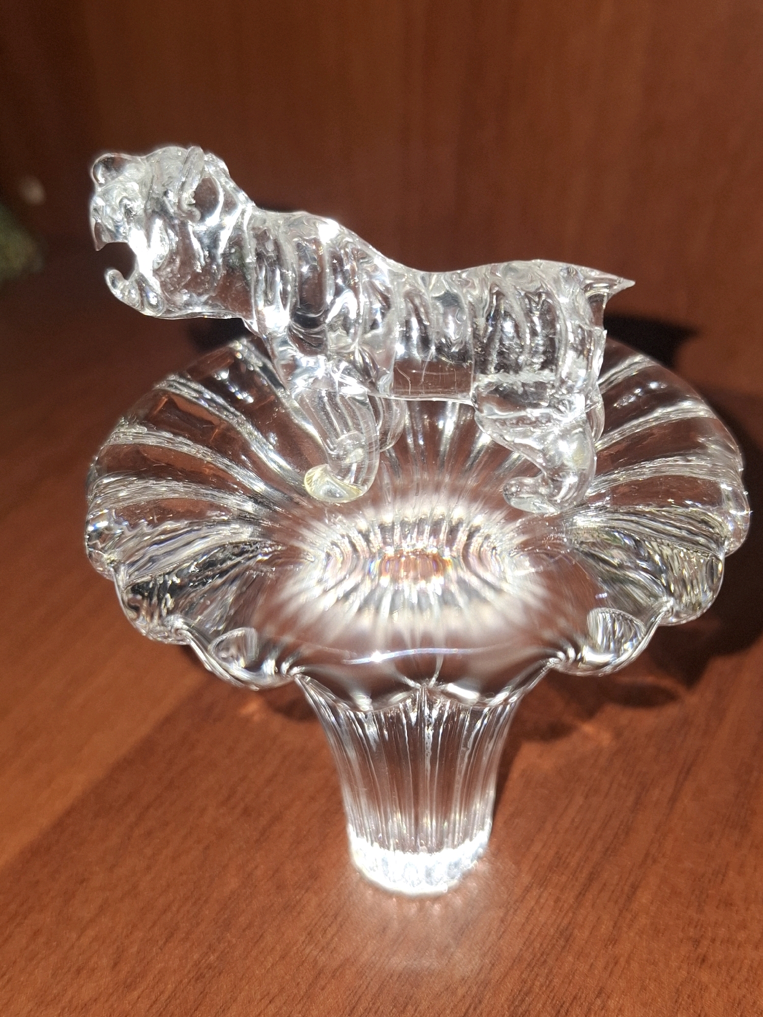 A clear glass figurine of a tiger is displayed on a fluted, glass pedestal. The tiger is positioned horizontally with its mouth open, appearing to roar or prowl. The pedestal is wide and shallow at the top, resembling a flower bloom, with vertical ridges extending downwards to a narrower base. The object is placed on a wooden surface with a visible grain, casting a soft shadow to the right. The background is a blurred, dark brown. There is no discernible location in the city of Almaty, Kazakhstan.