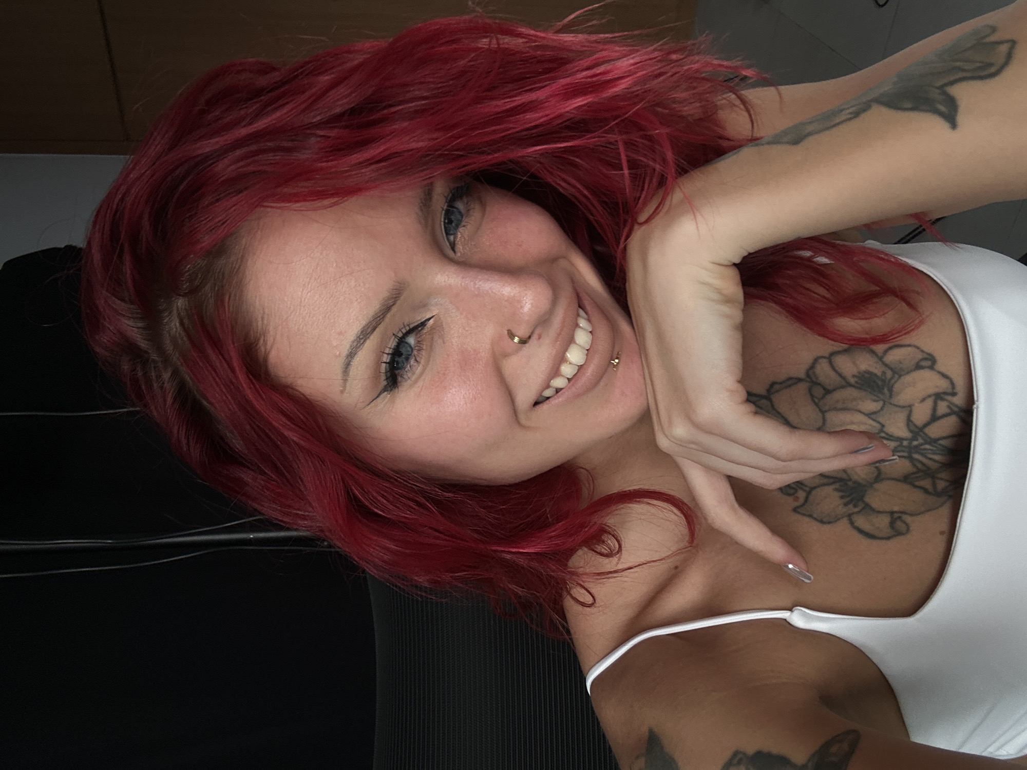 A fair-skinned woman with red hair and visible tattoos poses indoors. She has blue eyes, a nose piercing, and a lip piercing. She's wearing a white tank top. A floral tattoo is visible on her upper chest. Other tattoos are visible on her arms. The background is a mix of dark and light, with furniture or other objects partially visible. The location, North Kuta, Indonesia, is not visually determinable from the image.