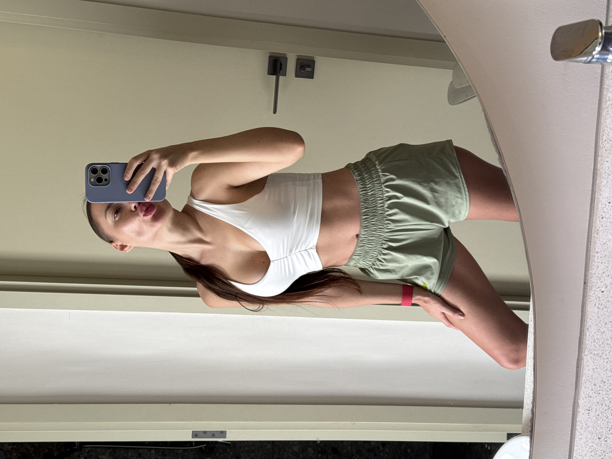 A woman is taking a selfie in a bathroom mirror. She is wearing a white sports bra and green shorts. She is holding a blue iPhone. The bathroom has light beige walls, dark grey door handle hardware, and a white sink. The location is likely a residence, but the city, Choeng Thale, Thailand, cannot be confirmed from the image.
