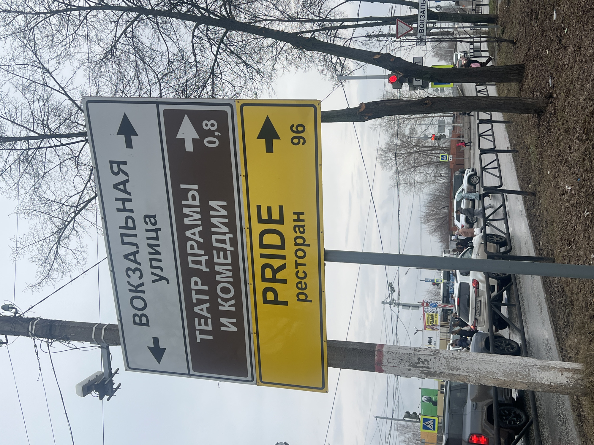 The image depicts a street signpost in Chaikovsky, Russia, with three direction signs. The top sign, with a gray background and black lettering, indicates that "Vokzalnaya Street" is to the left. The second sign, with a brown background and white lettering, indicates that the "Drama and Comedy Theater" is 0.8 units (presumably kilometers) straight ahead. The bottom sign, with a yellow background and black lettering, indicates "Pride Restaurant" is 96 units straight ahead. The signpost is made of concrete and appears weathered. In the background, there is a street with cars and pedestrians. There are bare trees along the roadside. A traffic light with a red light is visible. There is a pedestrian crossing signal visible. The weather appears overcast. The license plate of a white SUV reads 0479ck 152.