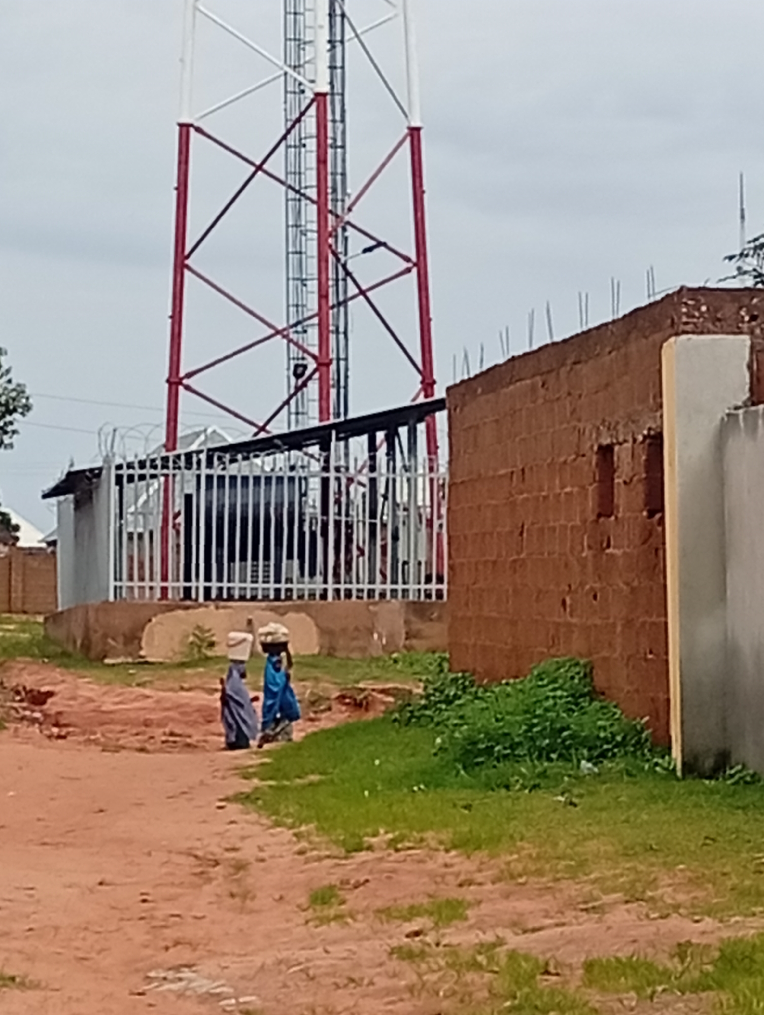 The image depicts a scene in Tumfure, Nigeria, featuring a prominent telecommunications tower in the background. The tower is characterized by its red and white lattice structure. In the foreground, a dirt path winds past a brick wall and some overgrown green vegetation. Two individuals are seen walking along the path, each carrying an object on their heads, likely for transport. One person wears a blue garment and the other a dark blue or black garment, partially obscured. A white metal fence encloses an area adjacent to the path, and behind it, partially visible, are some structures and equipment. The overall environment suggests a semi-urban or rural setting.