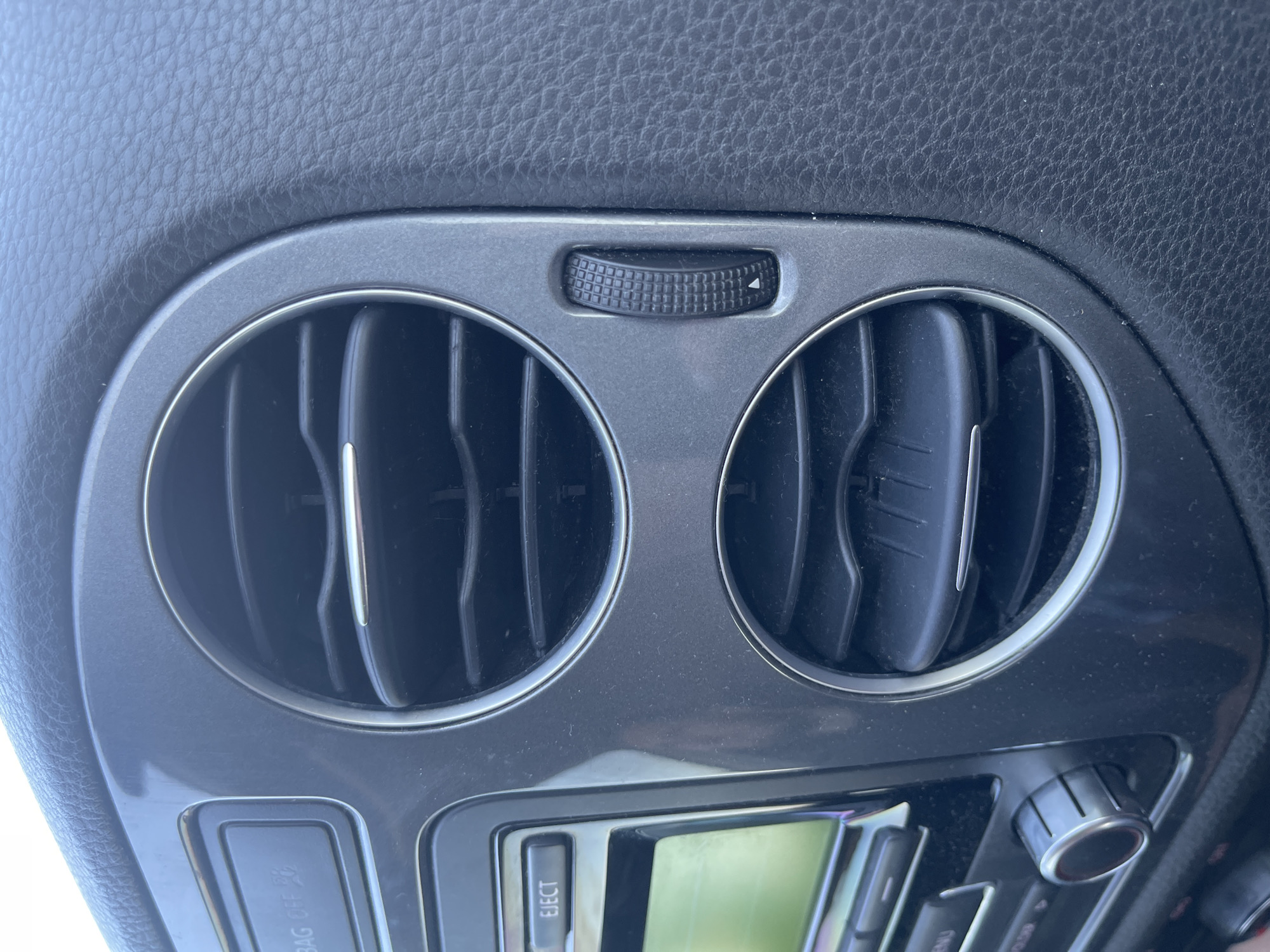 The image shows a close-up view of the interior of a car's dashboard, specifically focusing on a dual air vent and a part of the car's infotainment system. The air vents are circular with horizontal slats. To the left of the vents, a silver button labeled "EJECT" is visible above a screen. Below the screen, a rotary dial with silver accents is present. The dashboard is made of dark, textured material, likely plastic or leather. The lighting suggests an indoor or shaded environment, typical of a car interior. No people are visible in the image. The location of Novaky, Ukraine, is not identifiable from the visual elements in the image.
