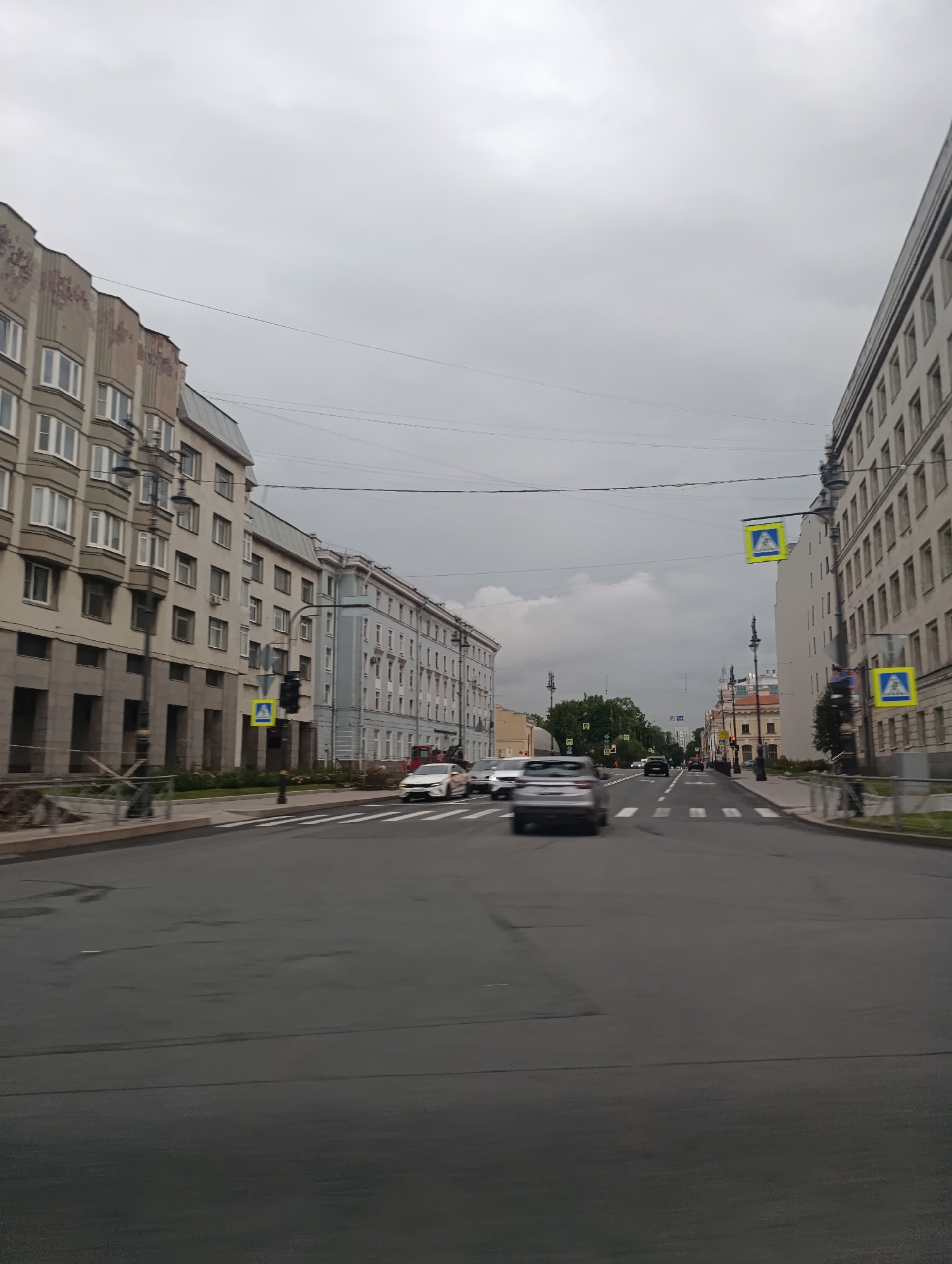 This image depicts a street scene in Saint Petersburg, Russia, under an overcast sky. The perspective is from a vehicle traveling down a wide, multi-lane road. On either side of the street are tall, multi-story buildings with numerous windows, characteristic of urban architecture. Some buildings show signs of wear and tear, with visible brickwork and possibly historical facades.
Several cars are visible on the road, some in motion, indicated by motion blur. Pedestrian crossing signs, marked with a yellow diamond and a white pedestrian symbol, are present at intersections, suggesting designated crosswalks. Traffic lights are also visible, though their current state (red, green, or off) is not clearly discernible. The road surface appears to be asphalt, with visible lane markings and some minor imperfections. Streetlights and electrical wires are strung overhead, contributing to the urban landscape. Vegetation, including trees, can be seen in the distance beyond the buildings. The overall atmosphere is that of a typical city street on a cloudy day.