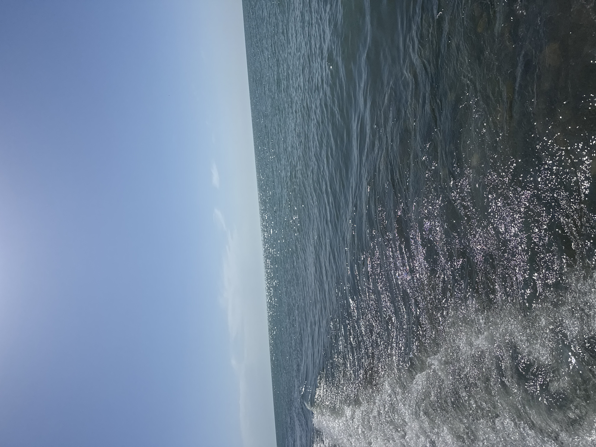 The image shows a body of water, likely an ocean or large lake, with a clear blue sky above. The water appears to be turbulent, with waves and white foam visible at the bottom right. The sky is a light blue color with a faint sun on the left side. There are no people or other identifiable objects in the image. The exact location is unknown.