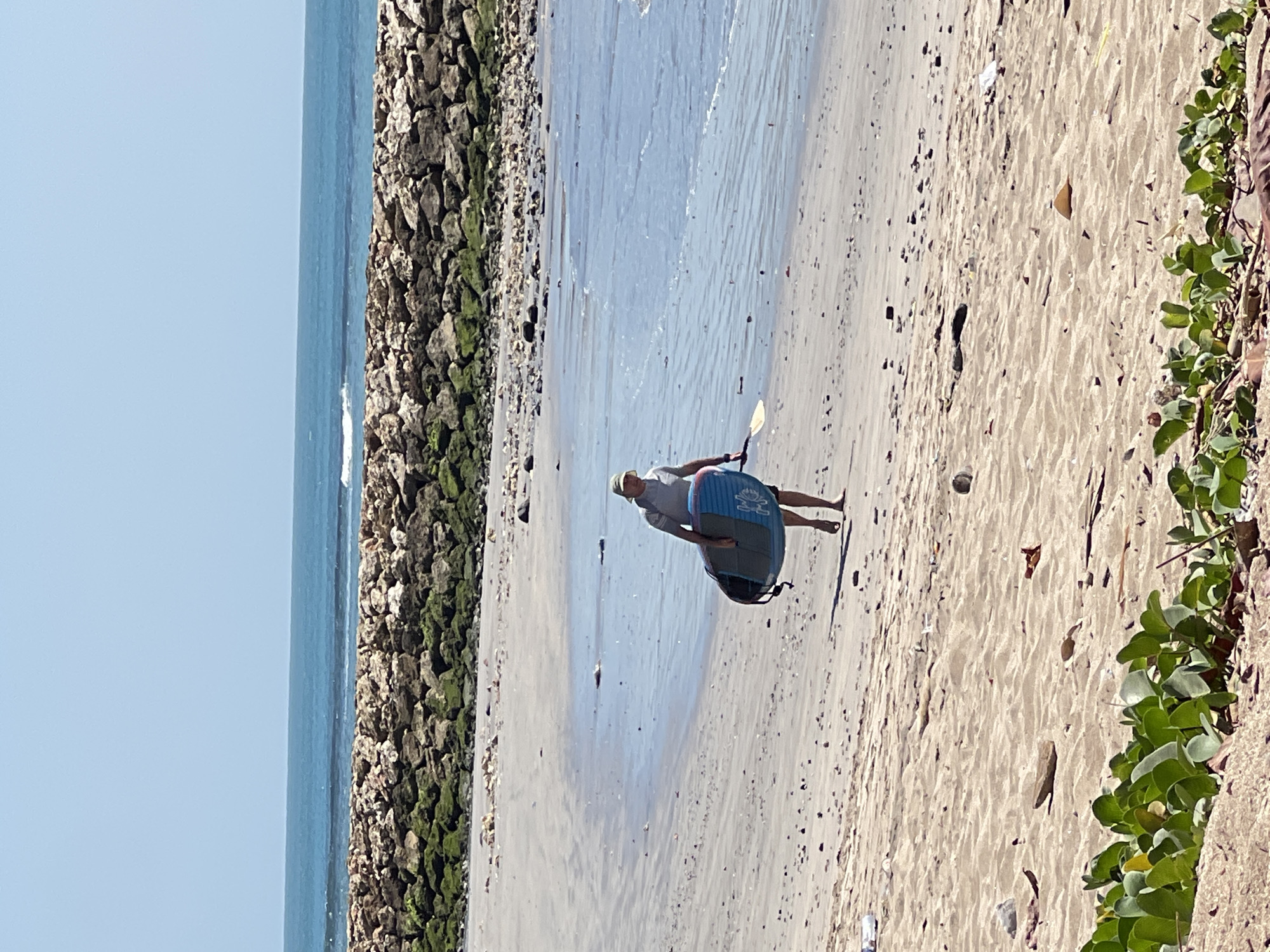 In Kuta, Indonesia, a person walks along a sandy beach carrying a stand-up paddleboard and a paddle. The person is wearing a light-colored hat and a grey t-shirt. The beach is littered with debris, including plastic bottles, debris, and driftwood. In the background, a stone breakwater is visible, and beyond that, the ocean stretches to the horizon under a clear blue sky. The water is calm with small waves breaking near the shore. Green vegetation grows in the foreground.