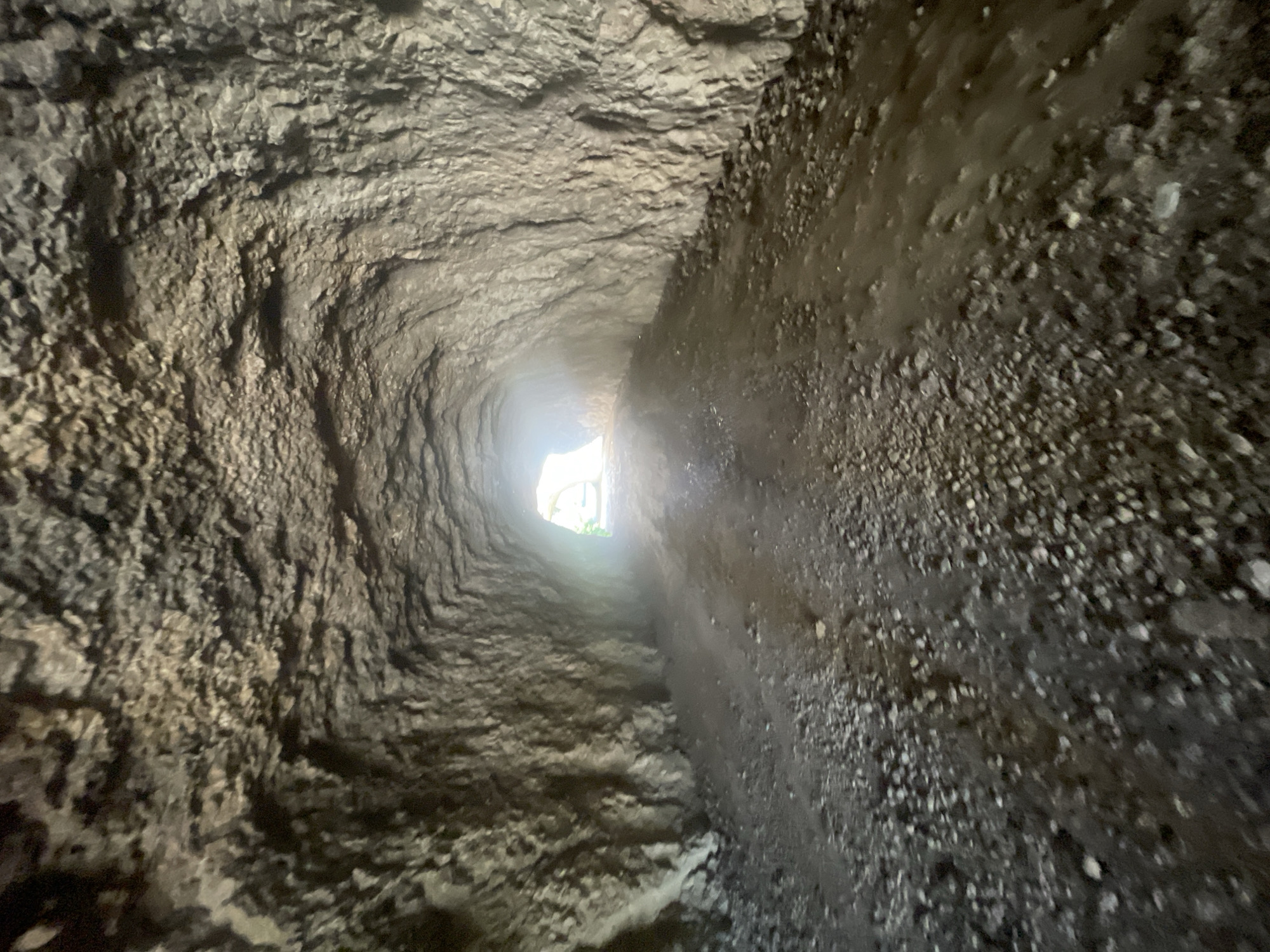 The image shows a narrow, rough-hewn tunnel or passage, likely underground or carved into rock. The walls are uneven and textured, with visible striations and a gritty appearance. The passage curves slightly, leading towards a bright light source at the end, suggesting an exit. The floor appears to be uneven and possibly damp, with small stones and dirt. The overall impression is one of an ancient or natural passage, possibly a cave, an old mine shaft, or an aqueduct. The lighting at the end of the tunnel is bright, hinting at daylight or a strong artificial light source outside. The scene conveys a sense of mystery and exploration, with the contrast between the dark, rough interior and the bright exterior. There is no text visible in the image.