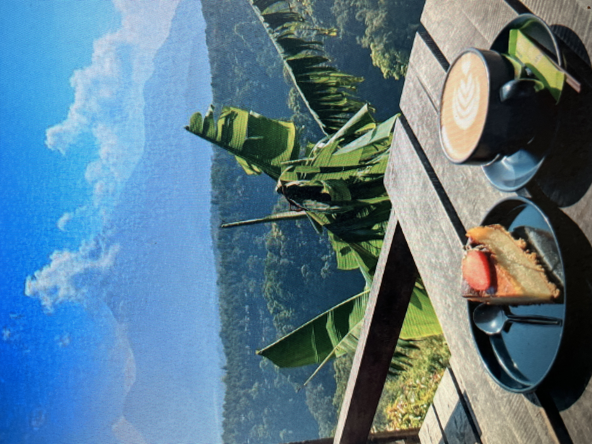 The image depicts a tranquil outdoor scene, likely from a cafe or restaurant with a scenic view. In the foreground, a slice of cheesecake topped with a strawberry slice and a spoon rests on a dark plate. Next to it, a dark mug filled with latte art sits on a saucer, with a green wrapper and a spoon peeking out from beneath it. The table is made of wooden planks, and in the background, lush green foliage and a distant mountain range are visible under a clear blue sky with scattered clouds. This scene is situated in Sejahtera, Indonesia.