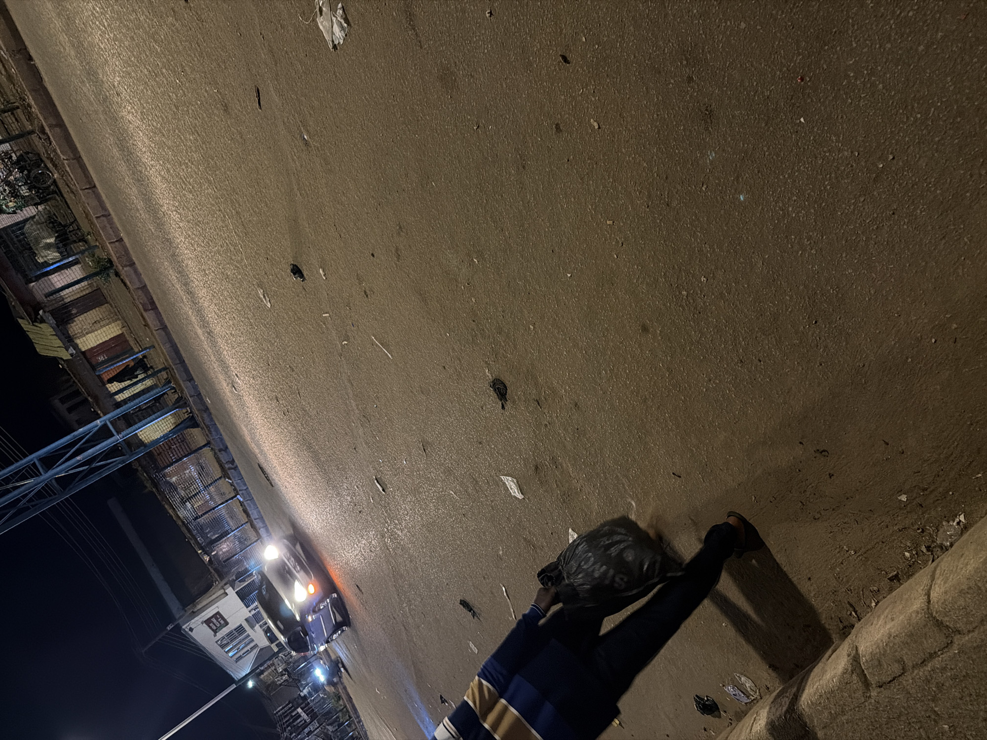 The image depicts a person carrying a black trash bag down a street at night in Bukuru, Nigeria. The person is wearing a dark long-sleeved shirt with yellow and white stripes on the sleeves, and appears to be wearing sandals. The trash bag is large and covers the person's head and shoulders, obscuring their face.
The scene is a paved street with a raised concrete curb on the right side of the image. There are various pieces of debris and litter scattered across the street, including what appear to be discarded papers and plastic wrappers. In the background to the left, there are buildings with lights on, suggesting an urban or semi-urban environment. A vehicle with its headlights on is also visible in the background, indicating ongoing activity.
The time of day is clearly night, as evidenced by the artificial lighting and the darkness of the sky. The weather appears to be clear. The presence of streetlights and building lights suggests it's an inhabited area. The overall impression is one of a routine task being carried out in a dimly lit urban setting. There is no visible text that can be clearly deciphered in the image.