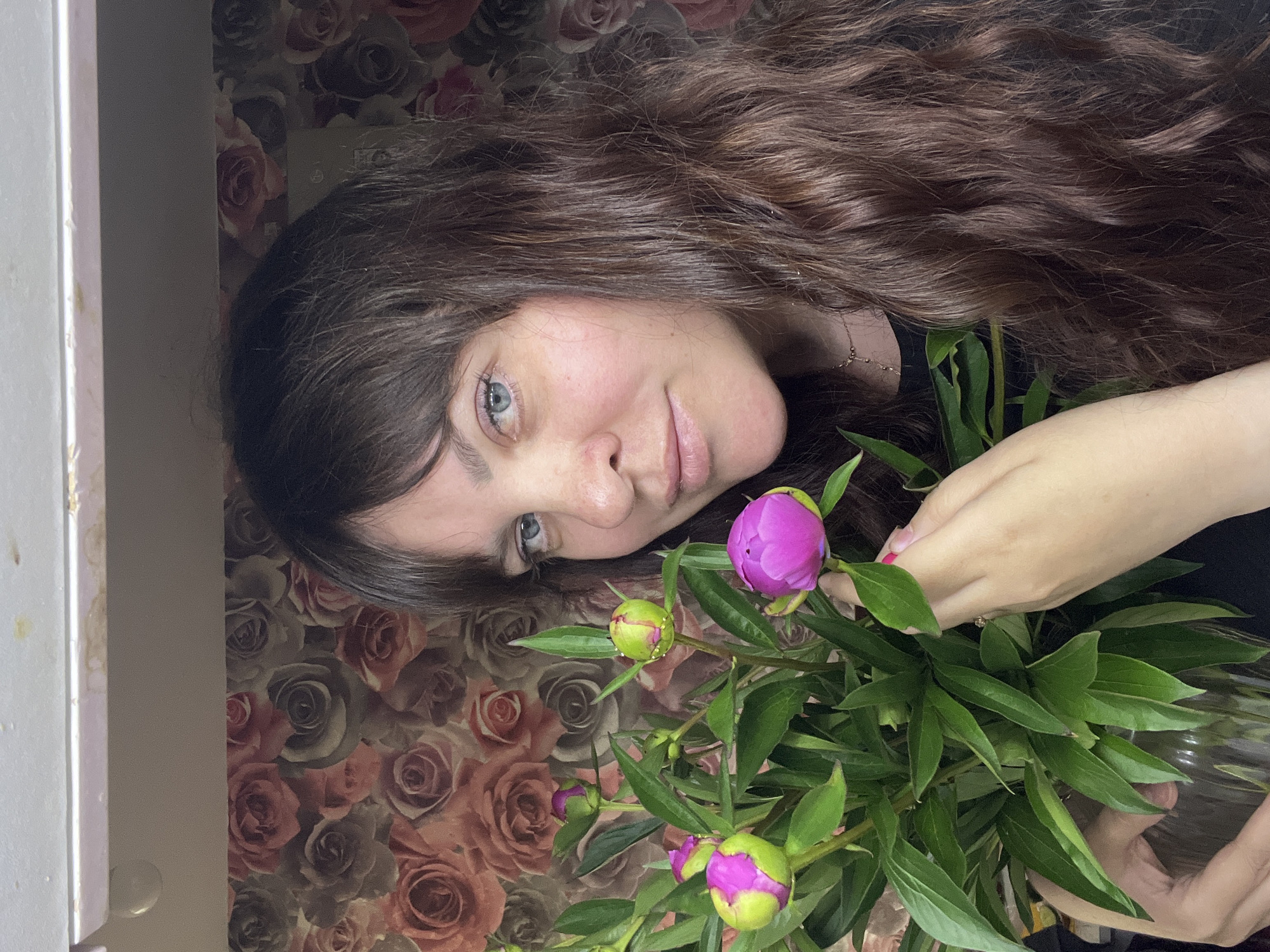 A medium shot shows a woman with long, dark brown, wavy hair and blue eyes posing with a bouquet of peonies. She is looking at the camera with a slight smile. The background is a floral wallpaper with roses and other flowers. The woman is wearing a black t-shirt and a delicate gold necklace. Her hands are holding the bouquet, which is in a clear glass vase. Several peony buds and one open flower, a vibrant pink, are visible. The lighting is soft and natural, suggesting an indoor setting. No specific location in Noviy, Russia can be identified from the visual information.