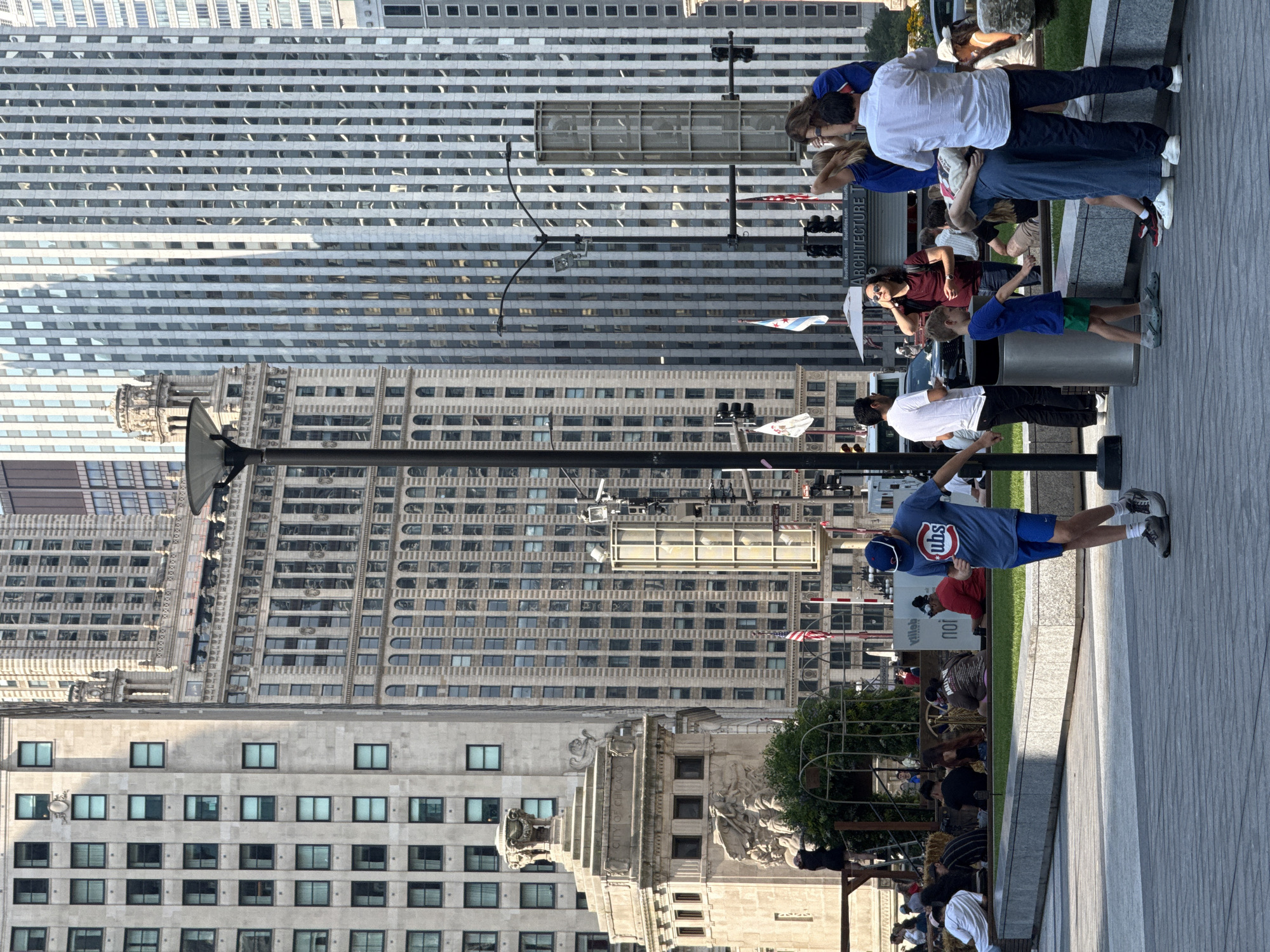 This is a daytime, eye-level, outdoor shot of a public plaza in Chicago, Illinois, United States of America. The plaza features a paved walkway and grassy areas, with several people present. In the foreground, a young boy in a blue t-shirt and green shorts is standing. To his right, a group of people are gathered, including a man in a white shirt and dark pants, and a woman in a blue top. Behind them, a woman in a denim jumpsuit is hugging another person.
The background is dominated by a cluster of tall, modern glass skyscrapers and older stone buildings, characteristic of Chicago's urban architecture. A lamppost with lights is visible in the mid-ground. Flags, including one with a blue and white design, are planted in the area. Signs for "Boat Rides" and "ARCHITECTURE TOURS" are also visible. Some people are seated on benches or the grass, and others are walking or standing. The overall scene depicts a lively urban gathering space.