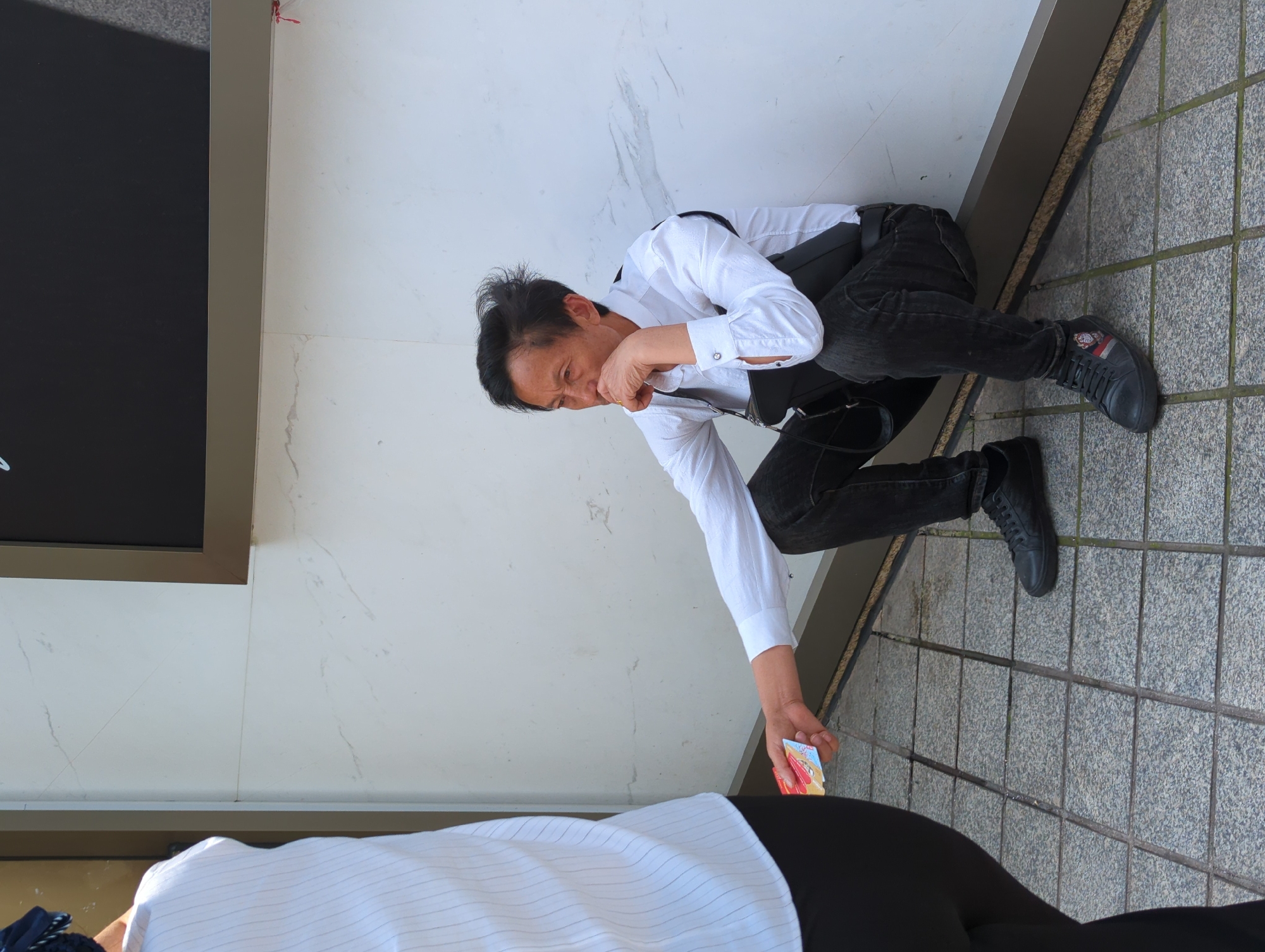 A man in a white shirt, black pants, and black shoes is crouching on a tiled surface in Yuzhong District, China. He is reaching for a small, colorful object on the ground, holding his face with his other hand, and looking down at the object. The background includes a white wall and a dark-colored sign. A small part of a person dressed in white is visible at the bottom of the frame.