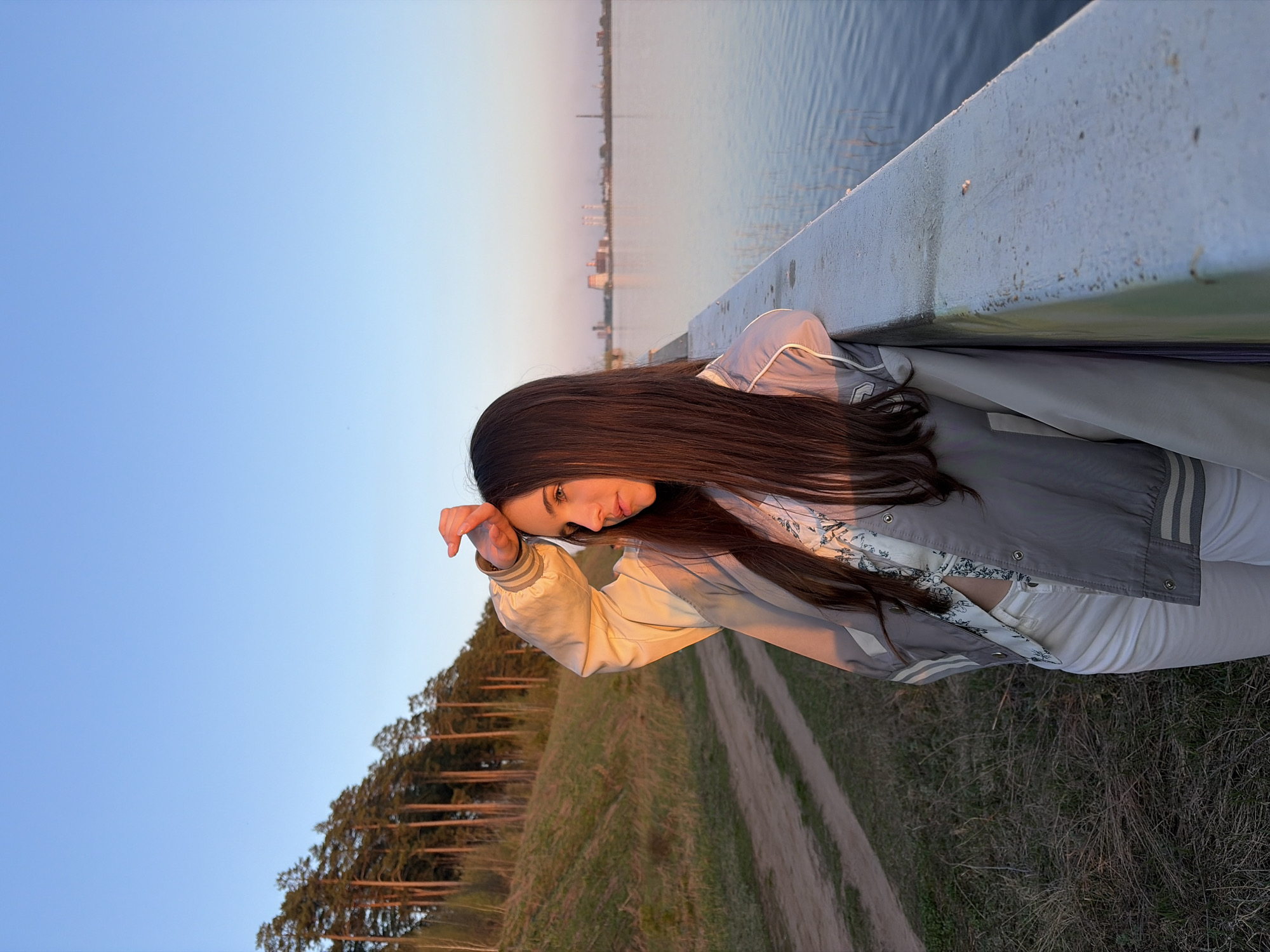 A young woman is depicted outdoors, seemingly resting her head on a railing or barrier. She is wearing a light-colored jacket over a patterned top, and white pants. Her dark hair falls over her shoulders. In the background, water and a cityscape can be seen, suggesting she might be near a river or lake. The lower portion of the image shows a grassy area with trees and a dirt path. This picture seems to be taken in Izhevsk, Russia.