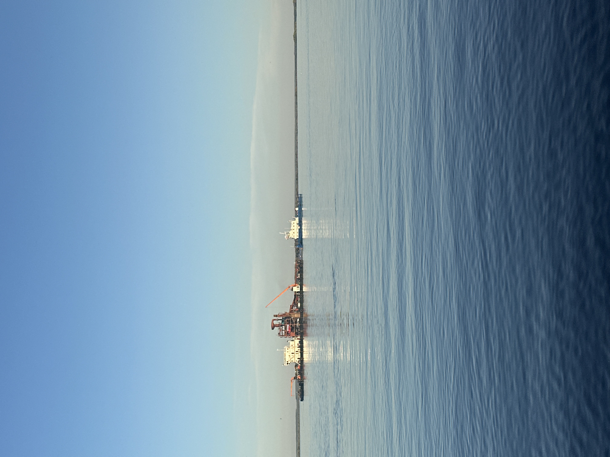 The image shows a body of water, possibly a wide river or lake, with a barge or construction platform in the distance. The sky is clear blue, suggesting a calm day. The water surface has slight ripples. The location is likely Soroch'i Gory, Russia.