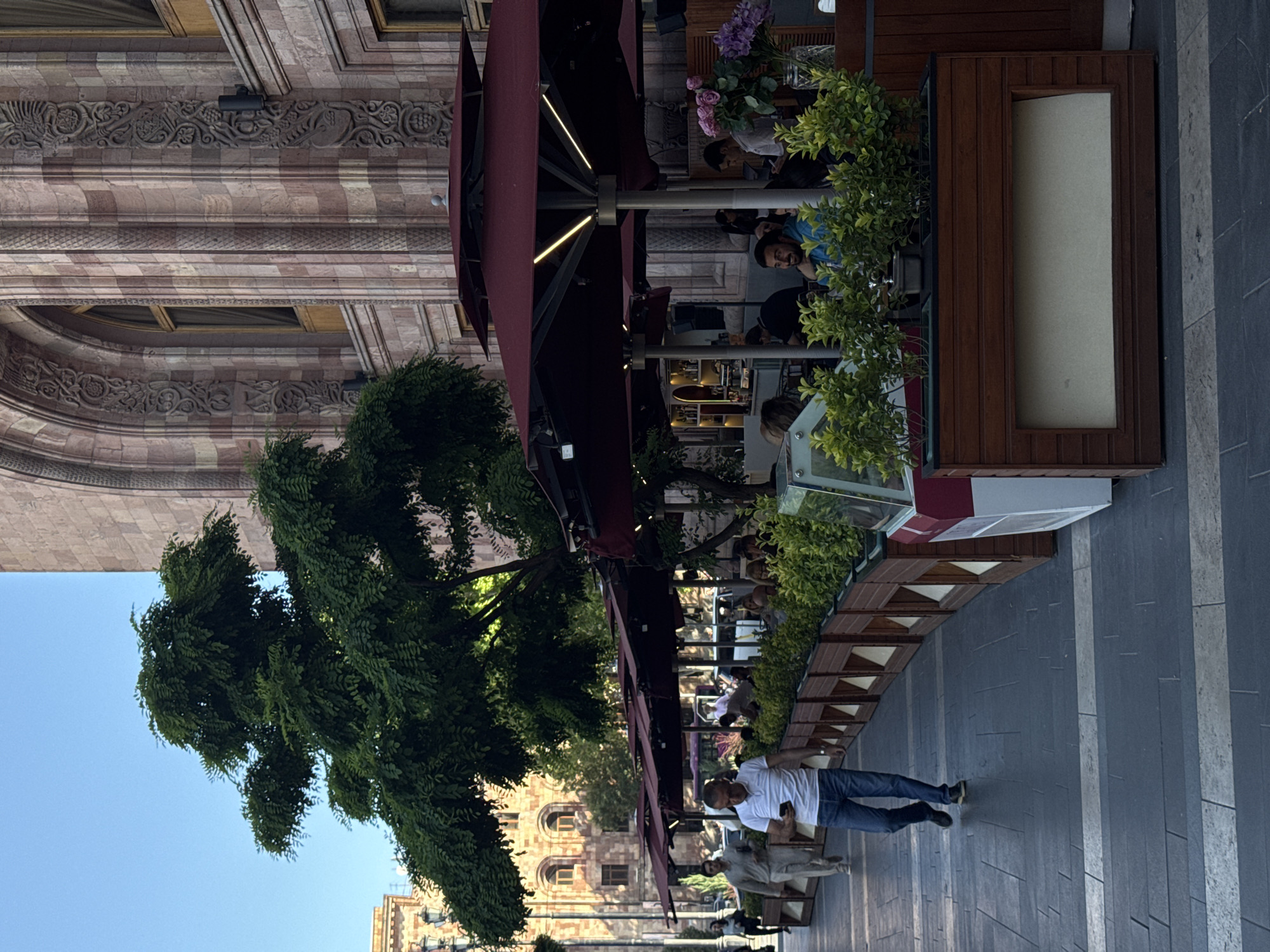 The image depicts a street scene in Yerevan, Armenia. The view includes pedestrians on a grey stone sidewalk, a cafe under red awnings, with people seated at tables, and a building with a patterned facade and arched windows. A mature tree is visible in the left foreground.