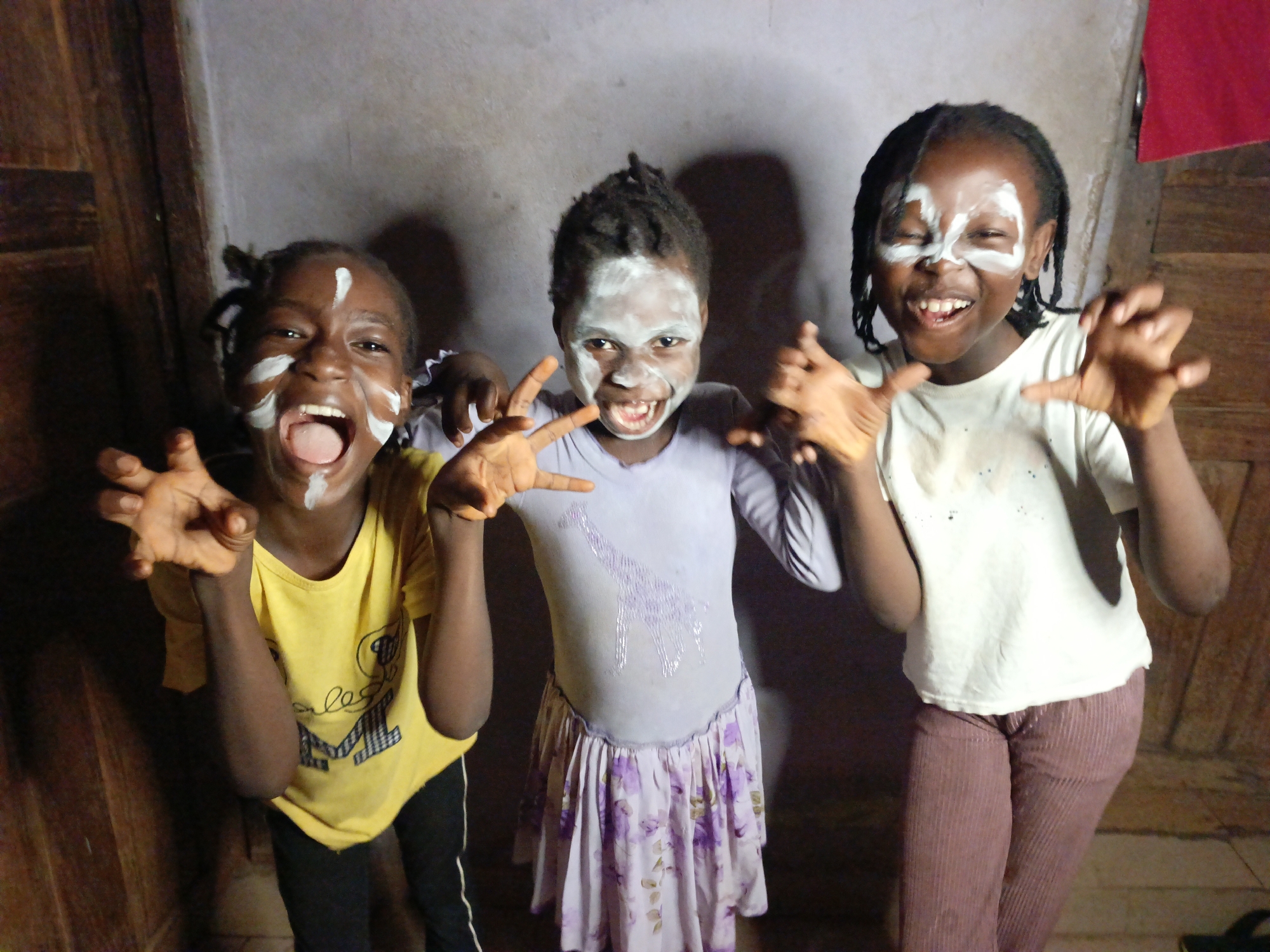 Three young children, likely girls, with dark skin and dark hair in braids or dreadlocks, are captured in a joyful and playful moment indoors. All three have white paint applied to their faces in stripes and patches, suggesting a festive or cultural activity. They are making exaggerated "clawing" hand gestures with splayed fingers, resembling animal paws.
The child on the left, wearing a yellow t-shirt with a visible "M" and some cursive text, and dark pants with white stripes, has her mouth wide open in a playful roar, exposing her tongue and teeth. The child in the middle is dressed in a light purple long-sleeved top featuring a glittery giraffe design and a light purple floral skirt. She holds her hands in a similar clawing gesture and displays a wide, happy grin. The child on the right, in a white t-shirt and reddish-brown ribbed pants, also has a wide-open mouth showing her teeth and makes a comparable hand gesture.
They are positioned in front of a rough, light-colored wall. To the left, a wooden door or panel is visible, and to the right, another wooden element and a hint of red fabric or object. The overall atmosphere is one of lighthearted fun and excitement, with all children displaying clear joy.