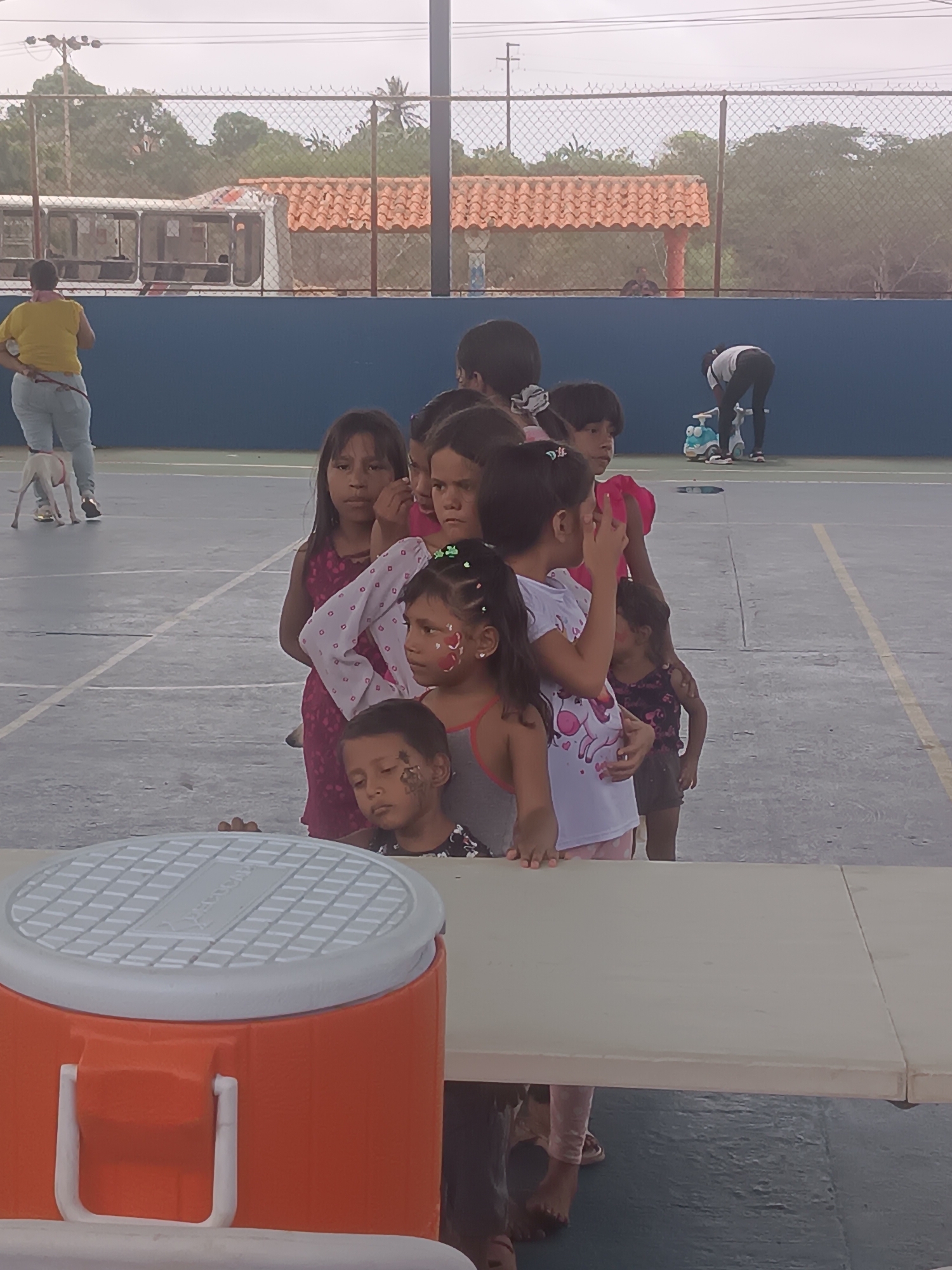 The image depicts an outdoor gathering in El Piache, Venezuela, under an overcast sky. In the immediate foreground, a white folding table is visible, with an orange and white plastic cooler placed on its left side. A group of approximately ten children, predominantly of Indigenous appearance and ranging in age from toddlers to early teenagers, are gathered behind the table. Some children have facial paint designs, specifically a girl with red and white marks on her cheek and a boy with a dark mark on his left cheek. They wear various casual clothing.
In the midground, to the left, an adult female wearing a yellow shirt and light-colored pants stands with her back to the camera, holding a leash attached to a white dog. To the right, another adult, dressed in a white shirt and dark pants, is bent over a small blue and white ride-on toy on a paved surface.
The background features a horizontal blue wall. Above this wall, a chain-link fence stretches across the scene, behind which are trees, dense foliage, and a structure with an orange-tiled roof. A white bus is partially visible behind the fence on the far left. Utility poles and overhead wires are also present in the upper left background. The ground is a dark grey paved surface with faint white and yellow markings.
