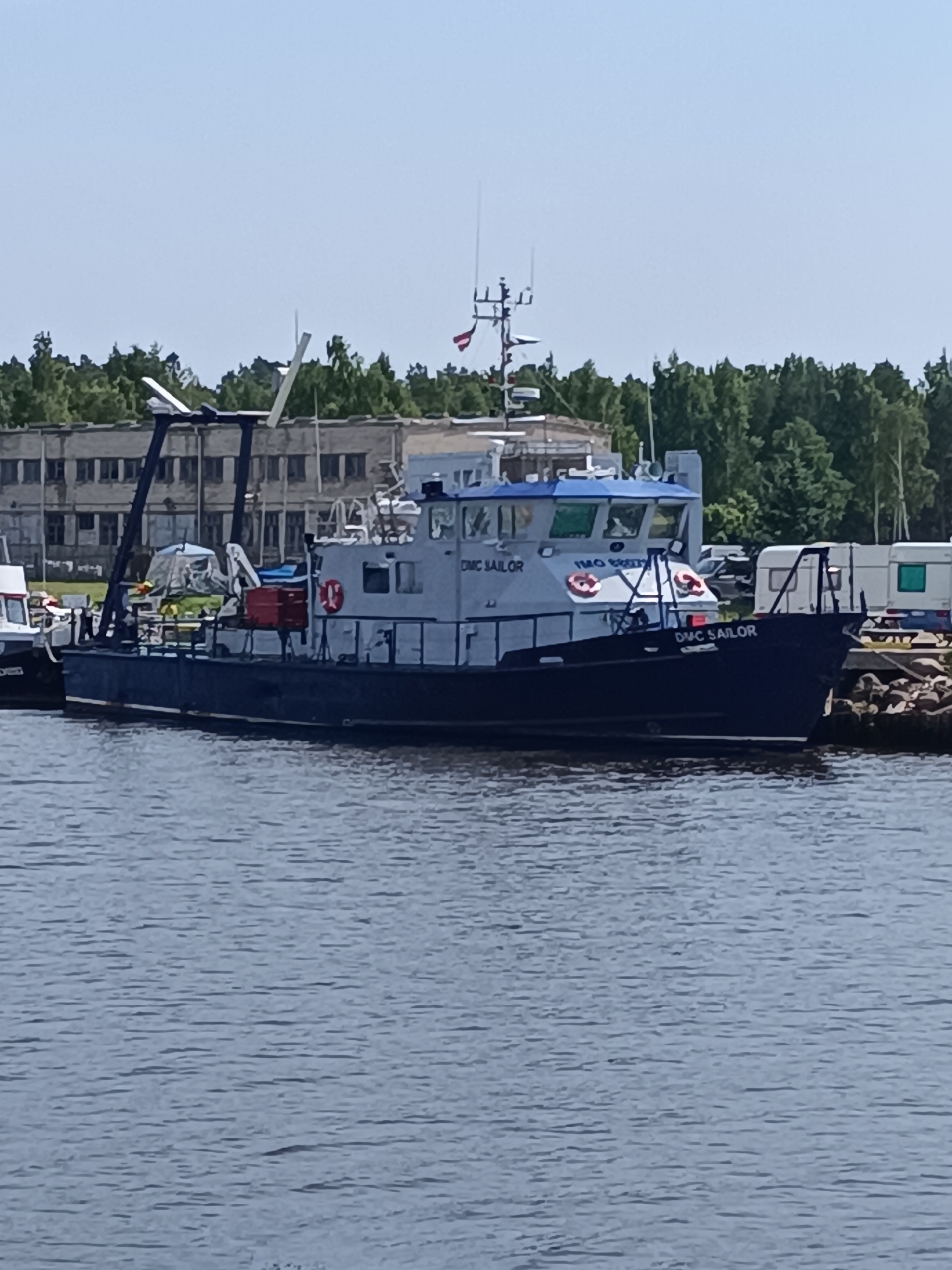 The image captures a vessel named "DMC SAILOR" docked in the harbor of Pāvilosta, Latvia. The boat, with a dark blue hull and a lighter blue superstructure, is positioned in the foreground, partially obscuring the water. The vessel is equipped with an A-frame structure at its stern and a smaller crane-like apparatus on its deck. Red lifebuoys are visible on the side of the superstructure, and the vessel's name is clearly displayed on its bow.
In the background, a low-rise, utilitarian building with multiple windows lines the shore. Behind the building, a dense line of green trees forms a natural backdrop. To the right of the vessel, several trailers are parked near a rocky embankment. The sky above is a clear, pale blue, suggesting fair weather. The water in the foreground exhibits a textured surface with numerous ripples, indicating a calm, yet active, harbor environment. No people are clearly visible in the image.