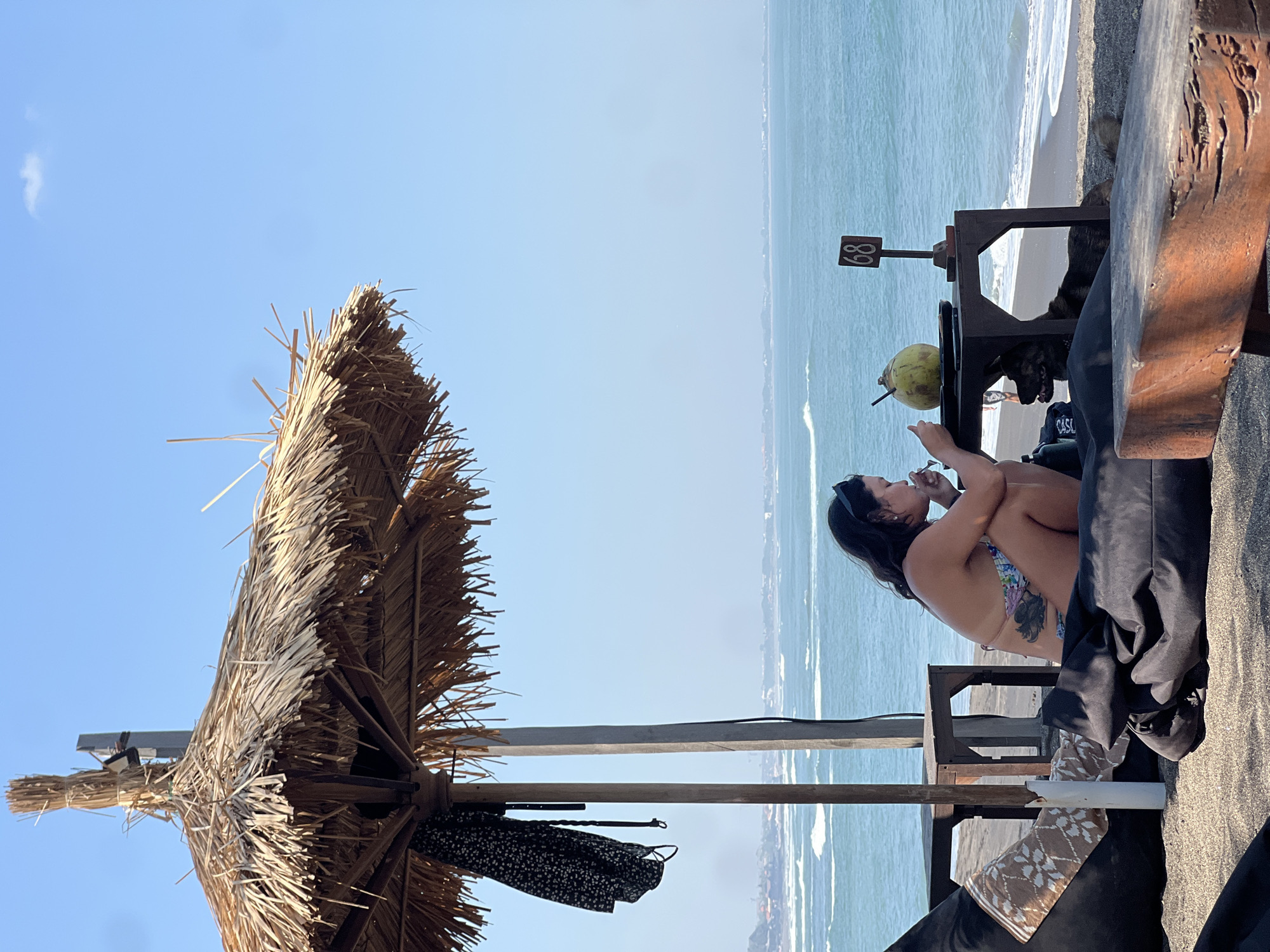 A woman sits on a beanbag chair under a thatched umbrella on a beach in Mengwi, Indonesia, eating from a bowl. A coconut with a straw and a table with a sign displaying the number "68" are to her right. A dog lies on the sand beneath the table. The ocean stretches out behind her, with waves breaking on the shore. The sky is clear blue with a small cloud. A black and white patterned dress hangs from the umbrella pole.