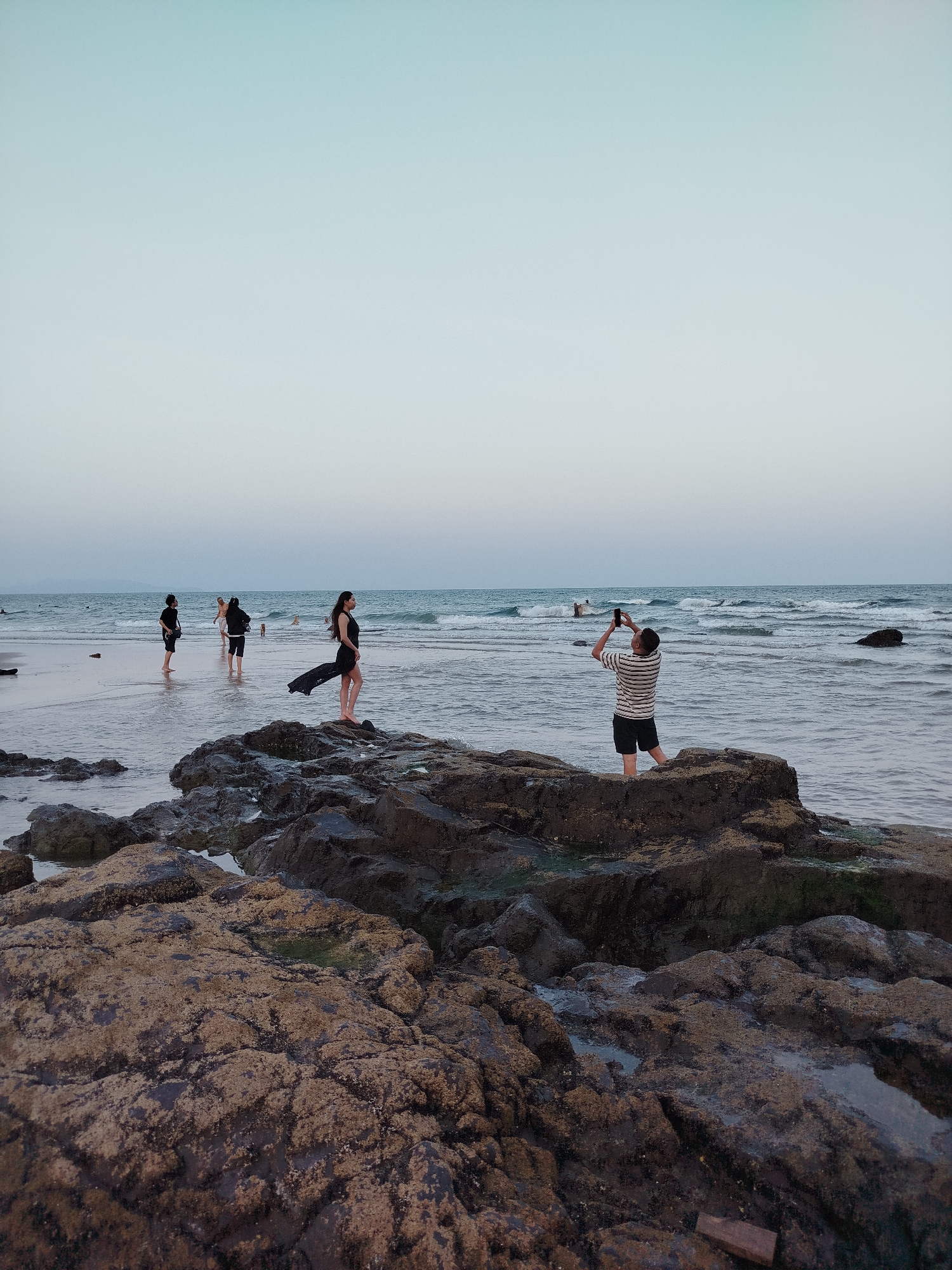 A wide, eye-level shot shows a beach scene under a pale sky. In the foreground, a cluster of dark, mossy rocks stretches across the lower portion of the frame. Behind the rocks, the ocean extends to the horizon.
Several people are positioned in the water. To the left, three individuals are wading in the shallow water near the shore. In the center of the frame, a woman wearing a black dress stands on the rocks. A man is seen photographing her. The location is Ho Chi Minh City, Vietnam.
