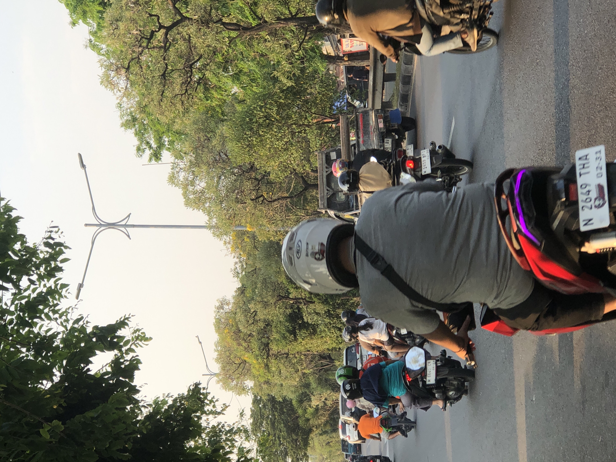 The image shows a street scene in Kludan, Indonesia, with a procession of motorcycles and a few cars. The perspective is from behind a motorcycle rider, looking forward. The road is paved and appears to be a multi-lane street, with traffic moving in the same direction. Lush green trees line both sides of the road, with some bearing yellow flowers. The sky is bright and overcast, suggesting it is daytime.
In the foreground, a person wearing a grey t-shirt and a white helmet is riding a motorcycle. The motorcycle has a red and black rear design with purple accent lighting. Behind them, other motorcyclists are visible, some with passengers. One person in the middle distance is wearing an orange t-shirt and a black helmet. Another rider is wearing a green helmet. Further ahead, a white car is visible, and beyond that, a small truck with a blue mud flap. The scene depicts typical urban traffic flow in Indonesia, characterized by a high volume of motorcycle usage. The license plates on some of the vehicles are partially visible, with one reading "N 2649 THA" and another "M 6640 CR." Streetlights with a modern design are also present along the road.