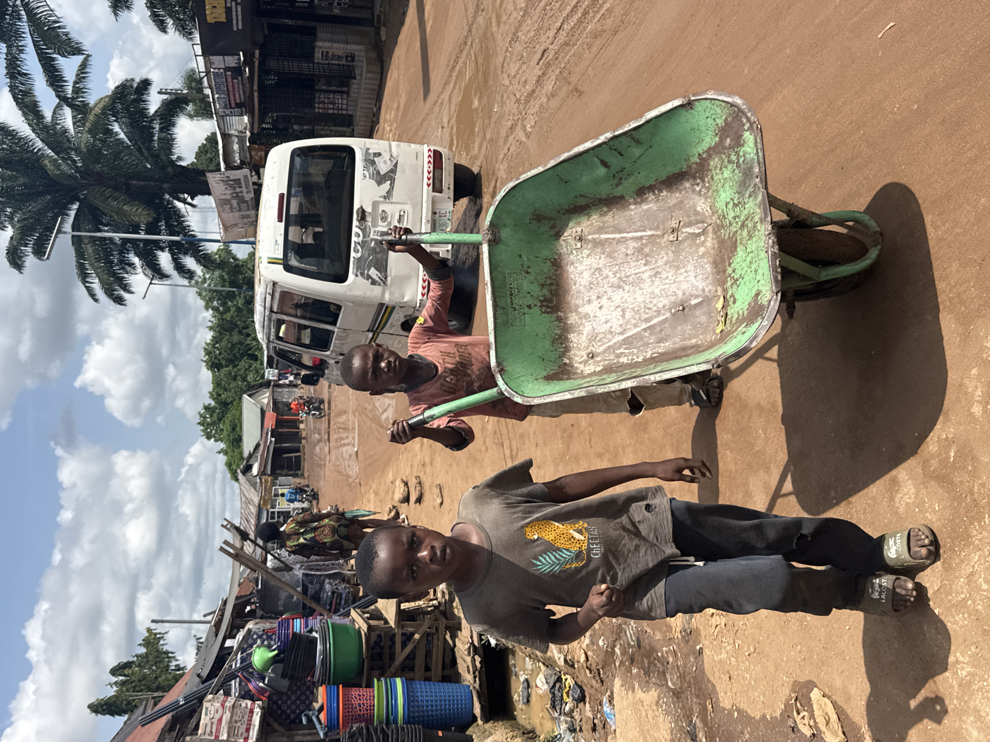 This image depicts a bustling street scene in Ihiagwa, Nigeria, under bright daylight. Two young boys are central to the composition. In the foreground, a boy, likely pre-teen, stands looking directly at the camera with a neutral expression. He is dressed in a grey t-shirt featuring a cheetah graphic and the word "CHEETAH," dark trousers, and olive green flip-flops with "LACOSTE" visibly printed on them.
Behind him, another boy, wearing a reddish-brown long-sleeved shirt, pushes a large, well-worn green wheelbarrow along a reddish-brown, unpaved road. The road surface shows areas of dry dirt mixed with muddy patches. A white passenger van or minibus, adorned with various stickers and a prominent red chevron graphic on its rear, is parked behind the boy pushing the wheelbarrow.
To the left of the foreground boy, market stalls are visible, stacked with colorful plastic buckets, basins, and other domestic items. Further into the background, simple buildings with corrugated iron roofs line the street. A large palm tree with dense fronds stands prominently against a clear blue sky dotted with white clouds, dominating the upper left portion of the image. The strong shadows cast by the subjects and objects indicate a sunny day, likely mid-morning or afternoon. The scene captures a moment of daily life in a vibrant, semi-urban or rural setting.