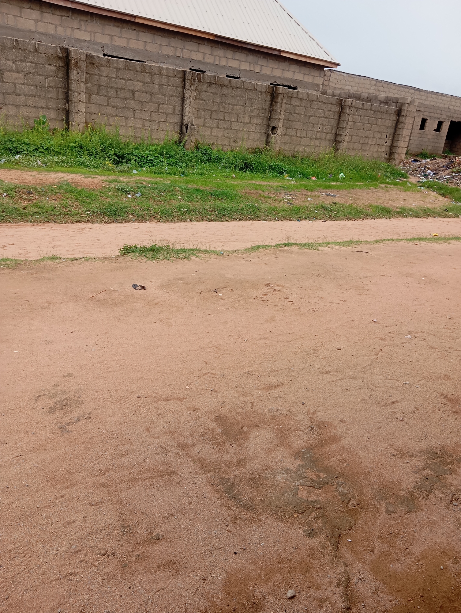 A low-angle, eye-level shot captures a dusty, reddish-brown dirt road in Jalingo, Nigeria, in front of a long, unfinished cinder block wall that forms the boundary of a building. The wall is approximately ten feet high and appears to be under construction, with sections of rebar visible at the top. To the left of the wall, a lush green patch of grass and weeds grows, interspersed with small pieces of white plastic debris. Beyond the grass, the facade of a partially constructed building is visible, featuring a light-colored metal roof and an upper section of beige painted blocks. To the far right of the scene, the wall continues and reveals dark openings that appear to be doorways or windows, with a pile of garbage and debris accumulated in the corner. The ground in the foreground is uneven and textured with footprints and small stones, indicating foot traffic. In the middle ground of the road, a discarded dark-colored shoe lies on the dirt. The sky is overcast with a pale gray hue.