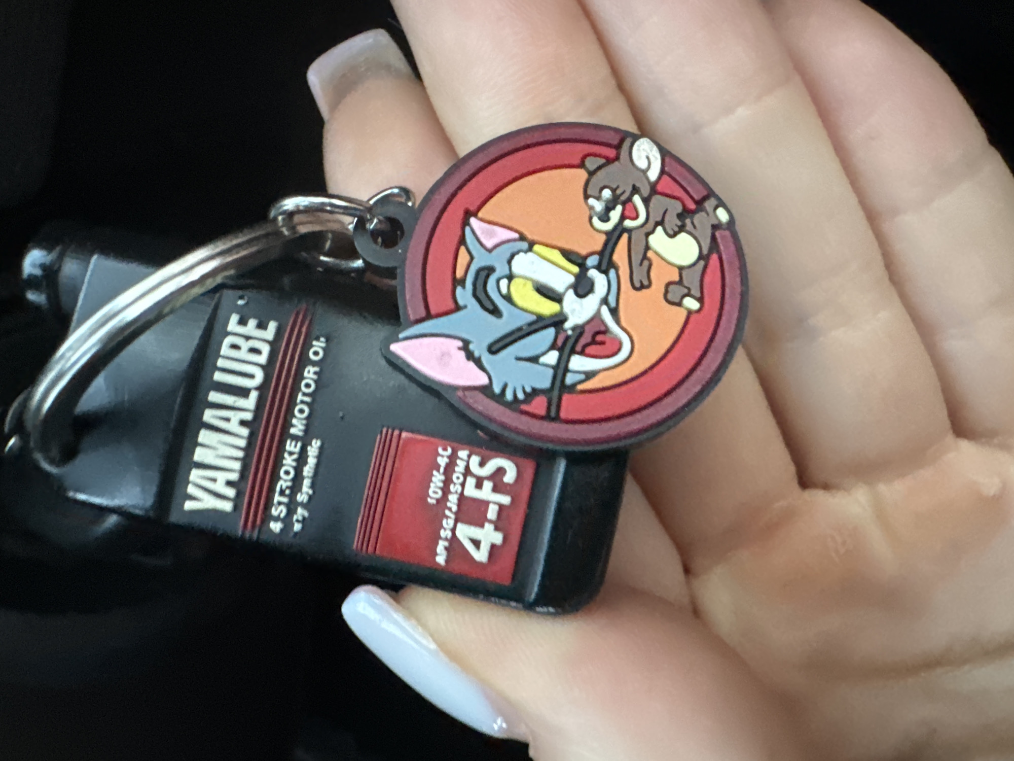A close-up, overhead shot shows a person's hand holding a keychain. The keychain features a miniature Yamaha oil bottle and a circular charm with cartoon characters Tom and Jerry. Tom is depicted in blue and yellow, with his mouth open and his tongue out, while Jerry is in brown, running. The background is dark and out of focus, suggesting an indoor setting. The image does not provide any information or visual cues related to the city of Murcia, Spain.