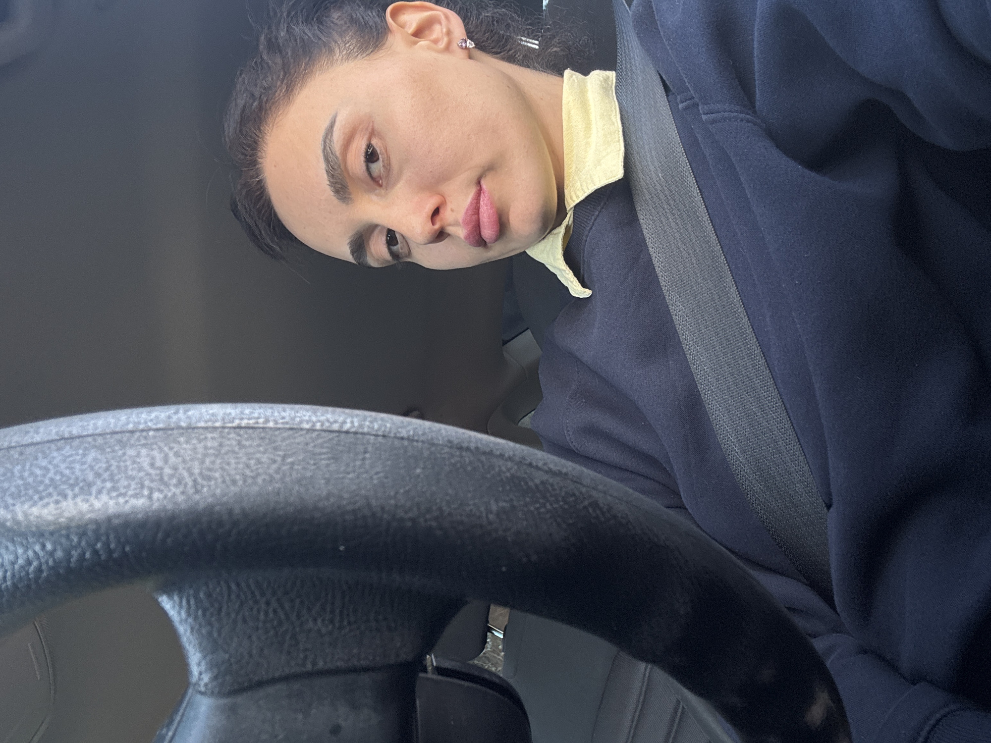 A woman wearing a dark blue sweater and a light yellow collared shirt is in the driver's seat of a car. She is wearing a seatbelt. A black steering wheel is in the foreground. The location appears to be Tbilisi, Georgia, based on visual characteristics associated with the region.