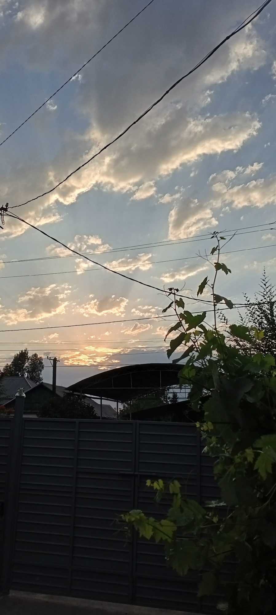 The image shows a sunset scene with power lines in the foreground, stretching across the frame from left to right. Behind the power lines, a sky filled with clouds is visible, with the sun setting behind a cluster of clouds, casting a warm glow. In the middle ground, there are buildings and a covered structure, possibly a carport or an open garage, with trees and foliage surrounding them. A dark, horizontal fence runs across the bottom of the image, obscuring the lower parts of the scene. The overall impression is a residential area at dusk, with the natural beauty of the sunset juxtaposed with man-made structures and infrastructure. The image is located in Sokuluk, Kyrgyzstan.