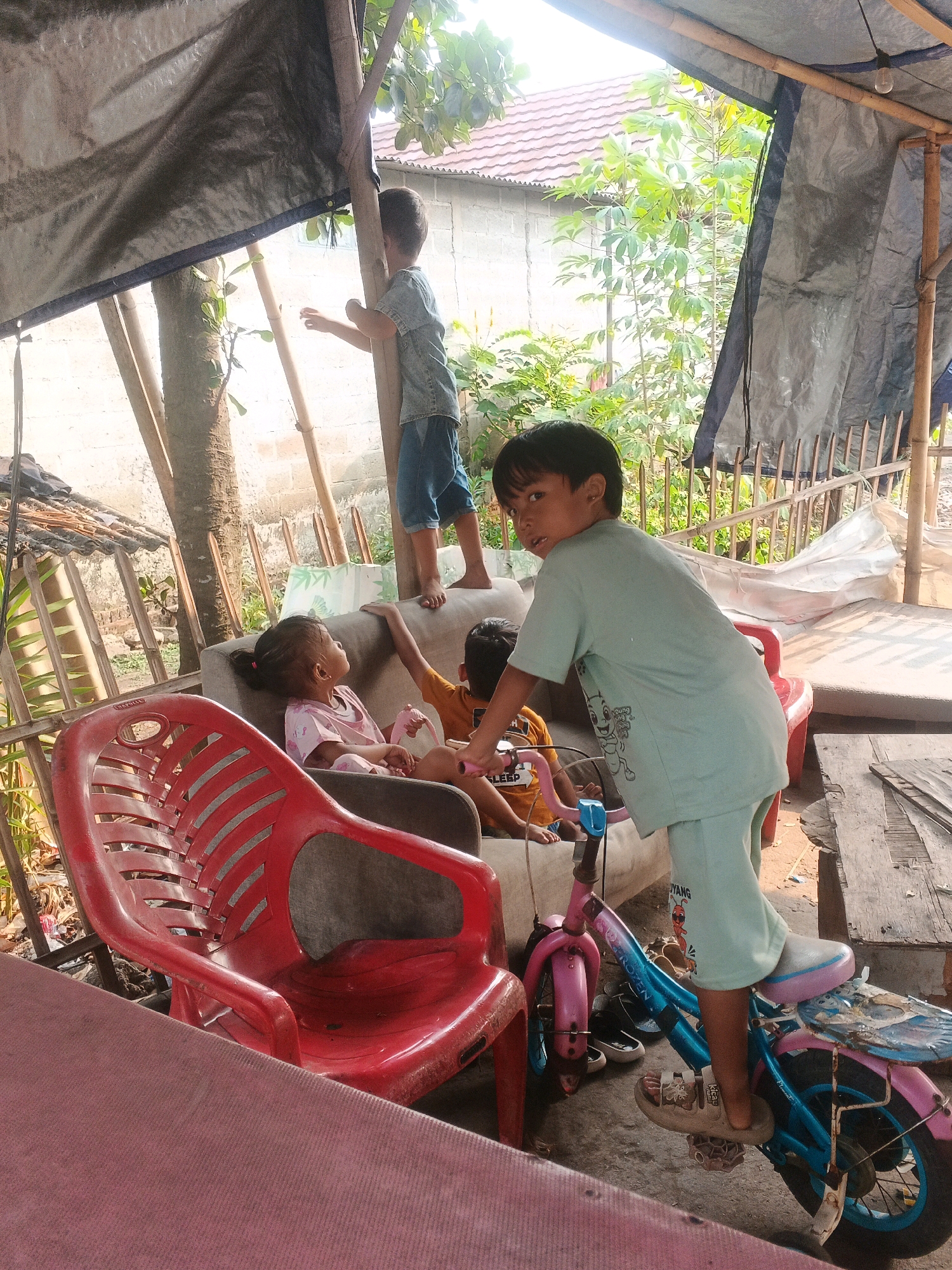 In Kadu, Indonesia, a group of children are gathered outdoors under a makeshift shelter. A young boy in a light blue t-shirt and shorts stands next to a pink and blue bicycle, looking towards the camera. Another boy, wearing a yellow shirt, sits on a couch, and a young girl in a pink shirt sits beside him. A third boy stands on top of the couch, looking away from the camera. The shelter is constructed with a dark tarp supported by bamboo poles. In the background, a white brick wall and lush green vegetation are visible. Red plastic chairs and a wooden table are also present in the scene. The overall environment suggests a simple, outdoor living space.