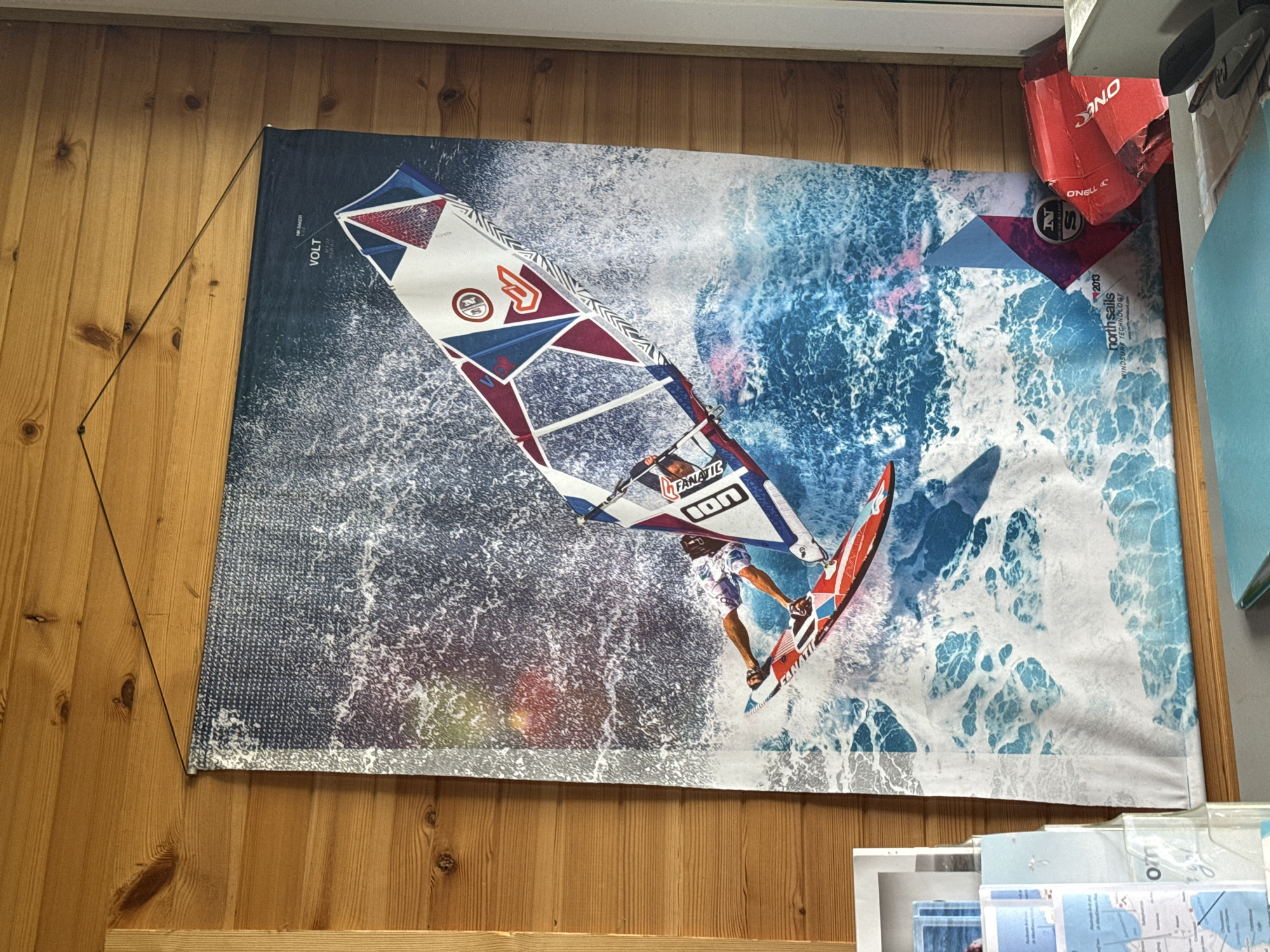 A banner featuring a windsurfer is displayed on a wall made of vertically aligned wooden planks. The banner showcases a windsurfer in action, with the sail prominently featuring "VOLT" and a prominent sail brand insignia. The waves surrounding the windsurfer are dynamically rendered with blues and whites, conveying motion. To the upper right of the banner are a red box labeled "O'NEILL" and some miscellaneous papers. Other papers are below the banner. The location is not stated.