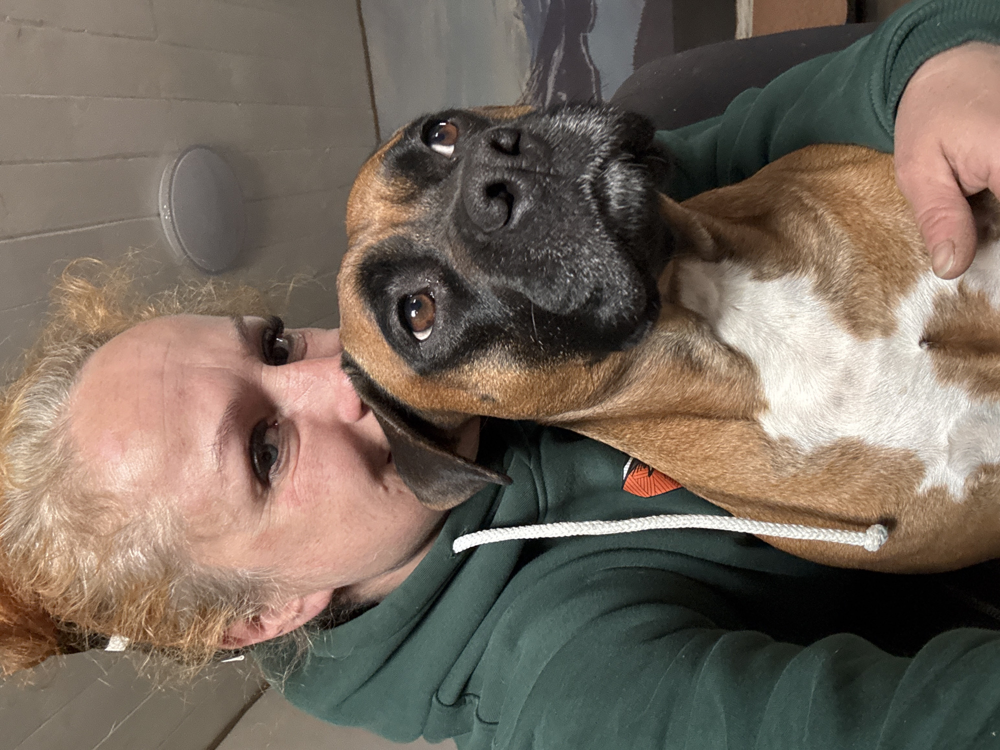 A selfie of a person with a Boxer dog is visible. The person has light-colored hair and is wearing a dark green hoodie. The Boxer dog is mostly brown with a black face and white markings on its chest. The dog is resting its head on the person. The background appears to be an interior setting. The location, Chaikovsky, Russia, is not discernible from the image.