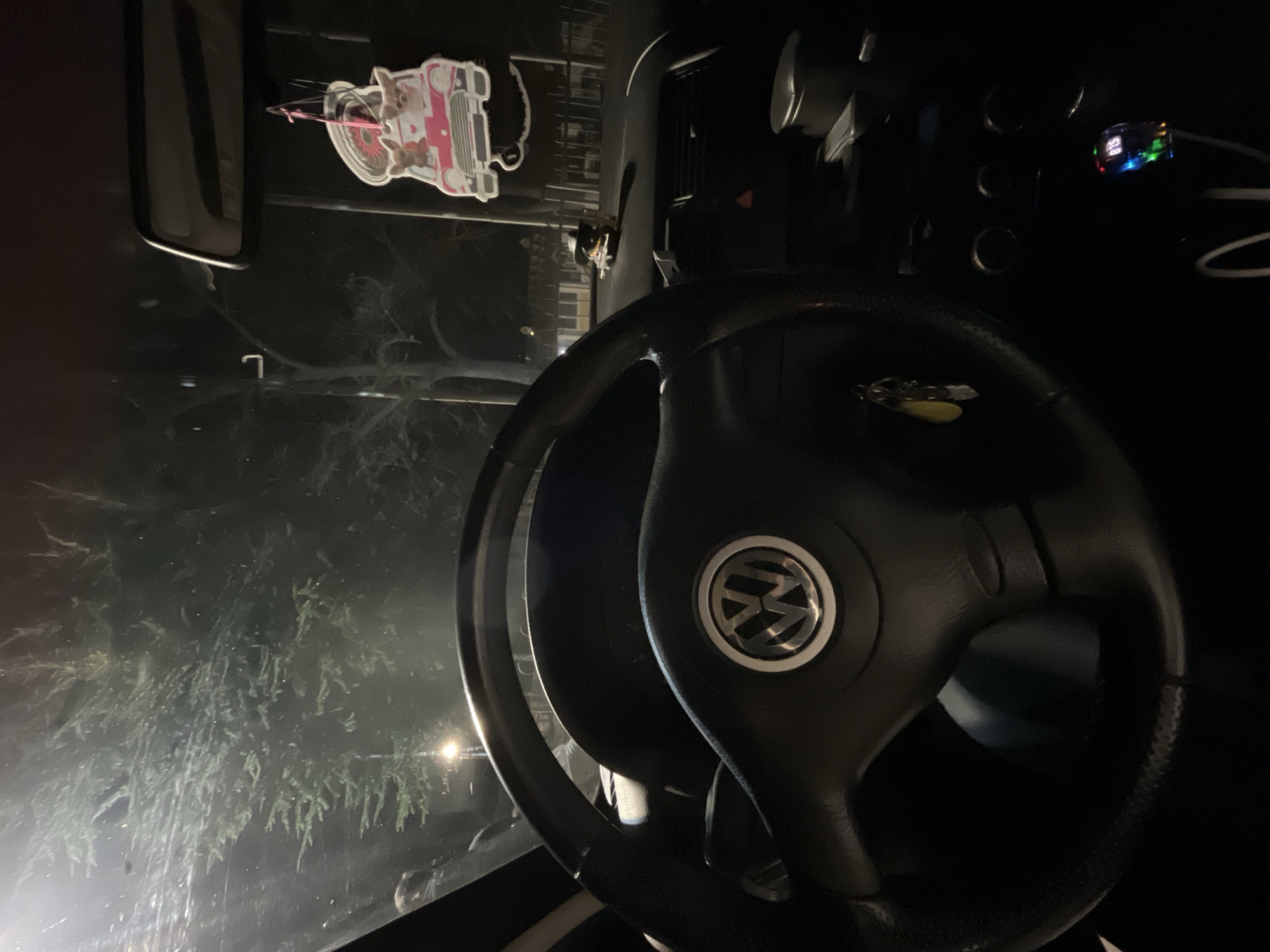 The interior of a car is visible. A black steering wheel with the Volkswagen logo is in the foreground. The dashboard contains several circular controls and a small display with glowing numbers. An air freshener shaped like a car is hanging from the rearview mirror. The windshield reveals a dark, indistinct scene outside, possibly a tree or bushes. The location appears to be Karavelovo, Bulgaria.