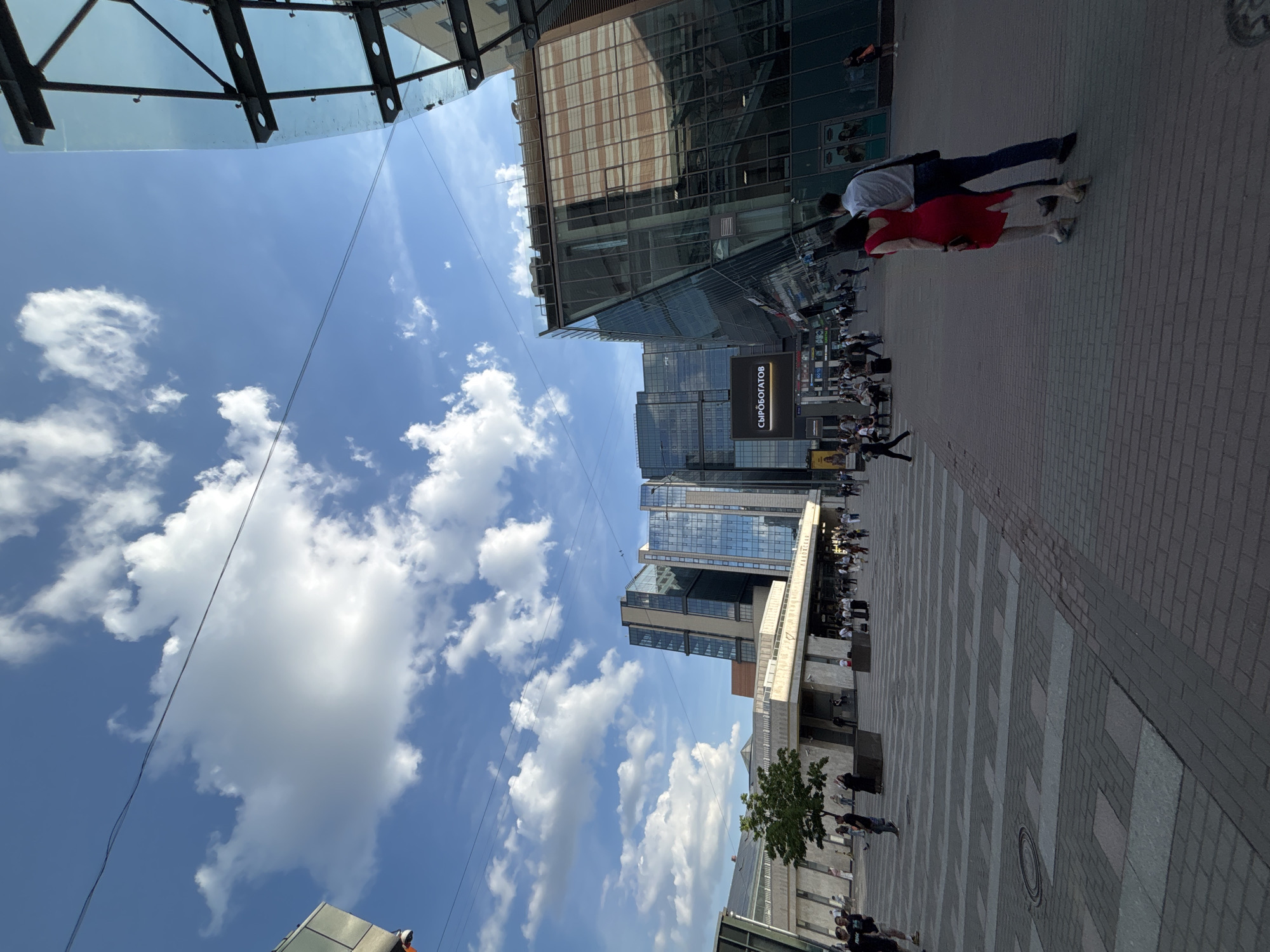 This image depicts a bustling urban scene in Saint Petersburg, Russia, specifically located at Vladimirskaya Square, featuring the Mayakovskaya metro station. The scene unfolds on a bright day under a clear blue sky interspersed with scattered white cumulus clouds.
In the foreground, a wide pedestrian plaza is paved with interlocking gray and beige rectangular pavers arranged in a linear pattern, with a circular manhole cover visible in the lower left. In the immediate foreground, a couple walks away from the viewer. The woman wears a red, sleeveless dress and light-colored high heels. The man beside her wears a light-colored collared shirt, dark trousers, and carries a dark shoulder bag.
To the left, a light-colored, multi-level concrete building, identified by a sign reading "МАЯКОВСКАЯ" (Mayakovskaya), serves as an entrance to the metro station. A small green tree stands directly in front of this building, and several dark, rectangular planters are positioned on the plaza. Numerous pedestrians are visible walking in and around the station entrance.
In the background and to the right, a series of modern, multi-story buildings with extensive glass facades dominate the skyline. These structures exhibit contemporary architectural designs, with reflective surfaces mirroring the sky and adjacent buildings. A large digital billboard on one of these buildings displays the text "СЫРОБОГАТОВ" in white lettering against a dark background. To the far right, a distinctive building features an angled glass facade supported by visible dark steel beams and frameworks. Thin overhead wires traverse the sky across the upper portion of the image. Numerous other pedestrians are scattered throughout the mid-ground and background, walking or standing within the urban environment.