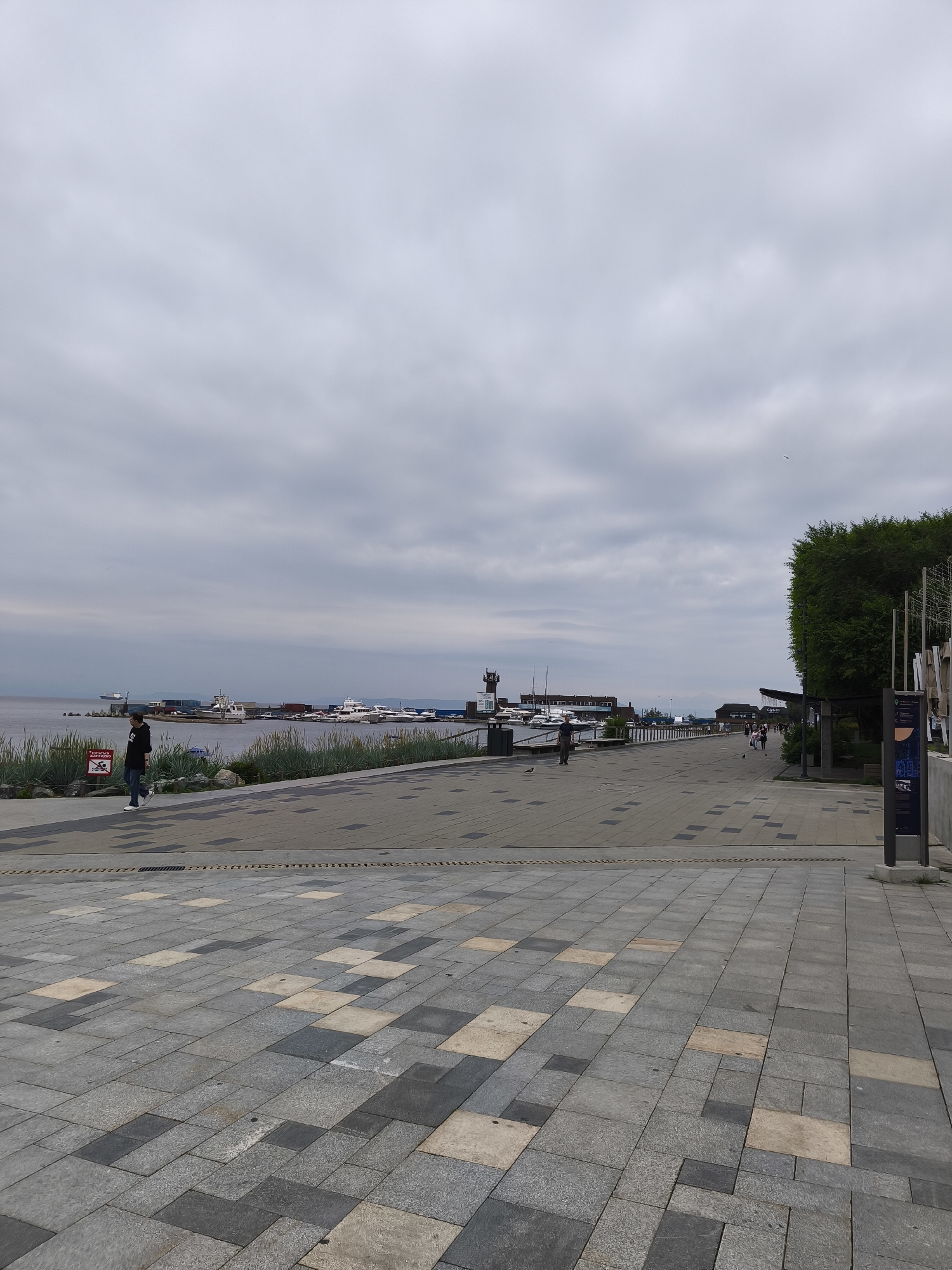 A wide, paved promenade in Vladivostok, Russia, runs along a harbor under an overcast sky. The promenade is paved with large, rectangular grey and beige tiles arranged in a pattern. To the left of the promenade, a body of water is visible, lined with lush green vegetation and scattered rocks. Several boats and yachts are docked in the harbor, with some buildings and a lighthouse visible in the distance. A single person is walking on the promenade, facing away from the camera. Further down the promenade, a small group of people are walking and standing. To the right of the promenade, a tall, dense green tree stands, casting a shadow on the pavement. A signage kiosk with an illustrated panel is also present on the right side of the promenade.