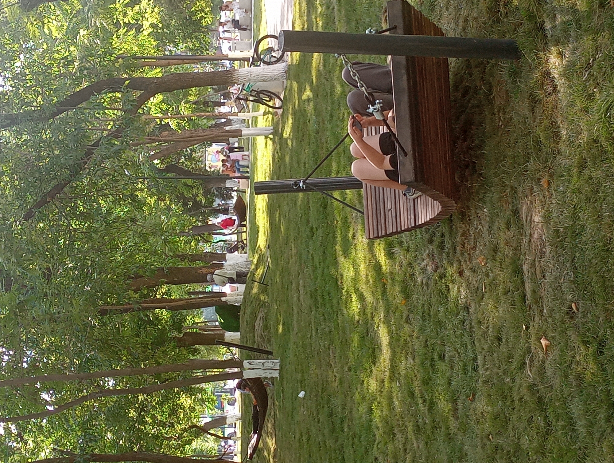 The image depicts a park scene in Shymkent, Kazakhstan, likely during the daytime with bright sunlight casting dappled shadows on the grass. A person is seated on a wooden bench, holding a phone and appearing engaged with it. They are wearing shorts and a t-shirt. A backpack rests on the bench beside them. The bench is positioned outdoors, with a grassy area and trees surrounding it. In the background, other people are visible in the distance, some sitting and others walking. A bicycle is propped up against a pole, suggesting recreational activity within the park. The overall atmosphere appears relaxed and casual, indicative of a typical day in a public park. The presence of trees and well-maintained grass suggests a well-kept public space. No visible text is present in the image.