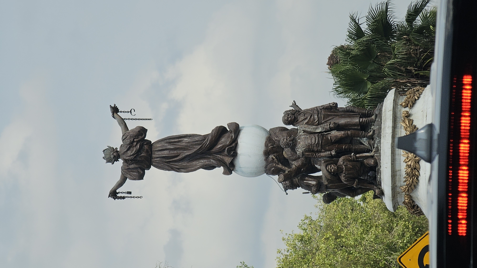 Here is a detailed description of the image:
The image features a statue of a woman standing on a globe, breaking chains held in her hands, which likely symbolizes liberation. The globe is supported by a group of male figures. The statue stands on a white pedestal adorned with golden leaf ornaments. Green trees and foliage surround the statue, and a palm tree is visible in the background. The sky is pale blue with light clouds. A portion of a vehicle, including a red taillight and a warning sign, is visible on the right side of the frame, suggesting the image was taken from a moving car.
Based on the statue's features and the setting, it is likely a photograph of the Altar to Liberty monument in Puebla, Mexico.