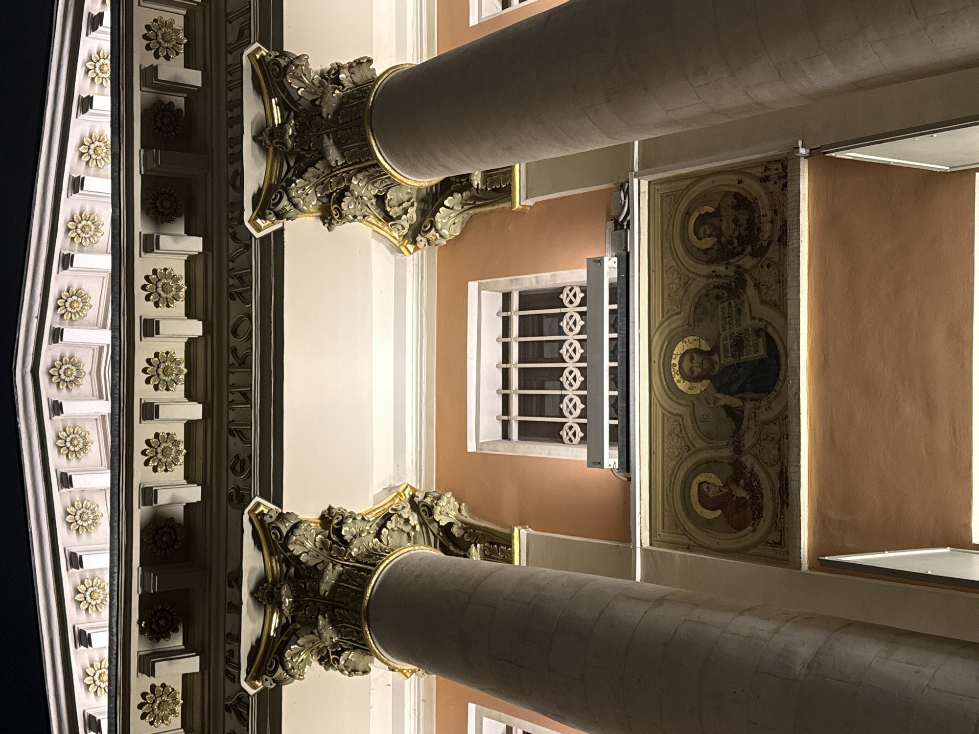 The image shows the interior architecture of a building, likely a church, in Moscow, Russia. Two large, cylindrical columns dominate the foreground, each with elaborately decorated capitals featuring floral and ornate designs in gold and dark green tones. A window with a white frame and decorative grillwork is visible in the background, set against an orange-toned wall. To the right of the window, an icon depicting religious figures is mounted on the wall. The overall perspective is from below, looking upwards towards the ceiling, highlighting the architectural details and decorations. The lighting is warm and suggests artificial illumination.