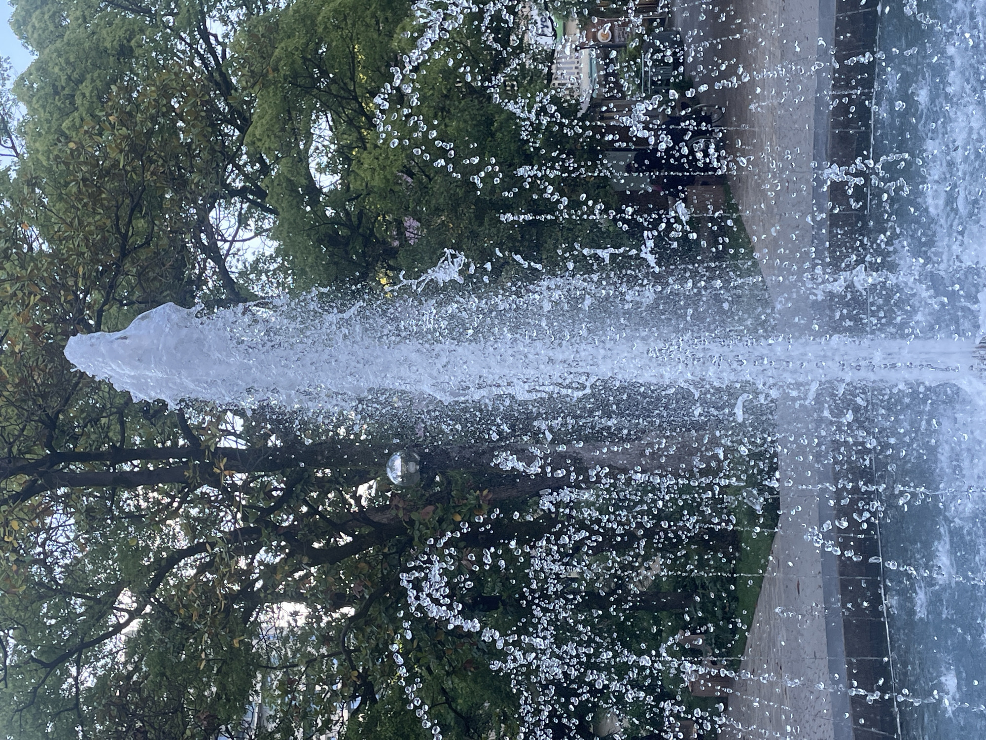 The image depicts a close-up view of a water fountain in a public outdoor space, likely a park or plaza, with a lush green tree canopy visible in the background. The fountain is actively spraying water upwards, creating a dynamic visual of water droplets in motion. A small, spherical, reflective object, possibly a disco ball or ornament, is suspended from one of the tree branches on the left side of the frame. The foreground shows the water basin of the fountain, filled with blue-tinted water. In the mid-ground and background, a paved pathway and further vegetation can be observed, along with hints of buildings or structures and some indistinct human figures, suggesting an urban environment. The presence of trees and a fountain indicates a potentially serene or recreational area within a city. The metadata indicates the image was taken in Sochi, Russia.