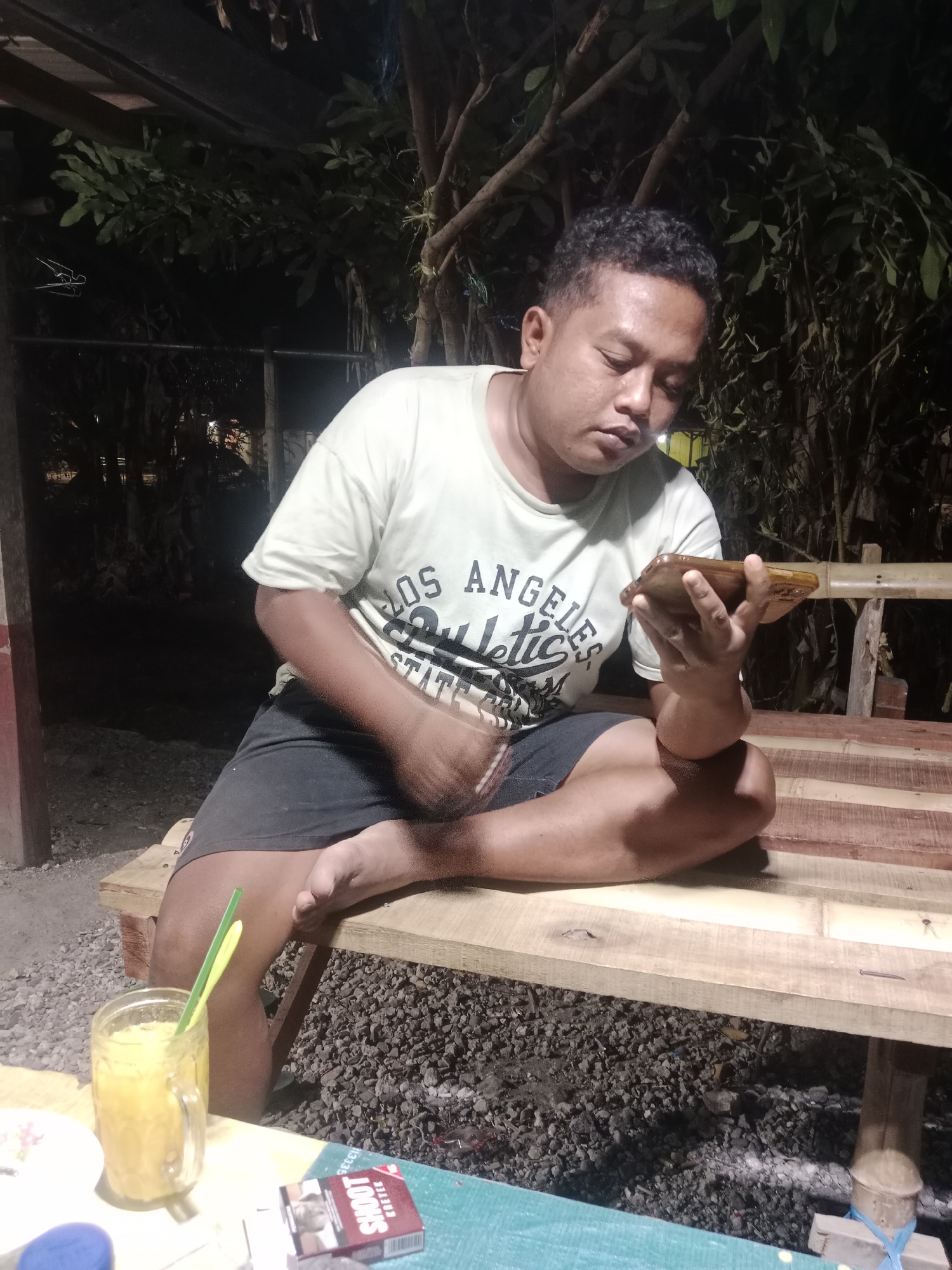 In Glagah Wangi, Indonesia, a man sits on a wooden bench, holding a mobile phone. He is wearing a light-colored t-shirt with lettering and dark shorts. In front of him, on a table, is a glass of orange juice with two straws and a pack of cigarettes. The scene appears to be outdoors, with trees and foliage in the background, suggesting a natural or semi-rural setting. The lighting indicates it is likely evening or night.