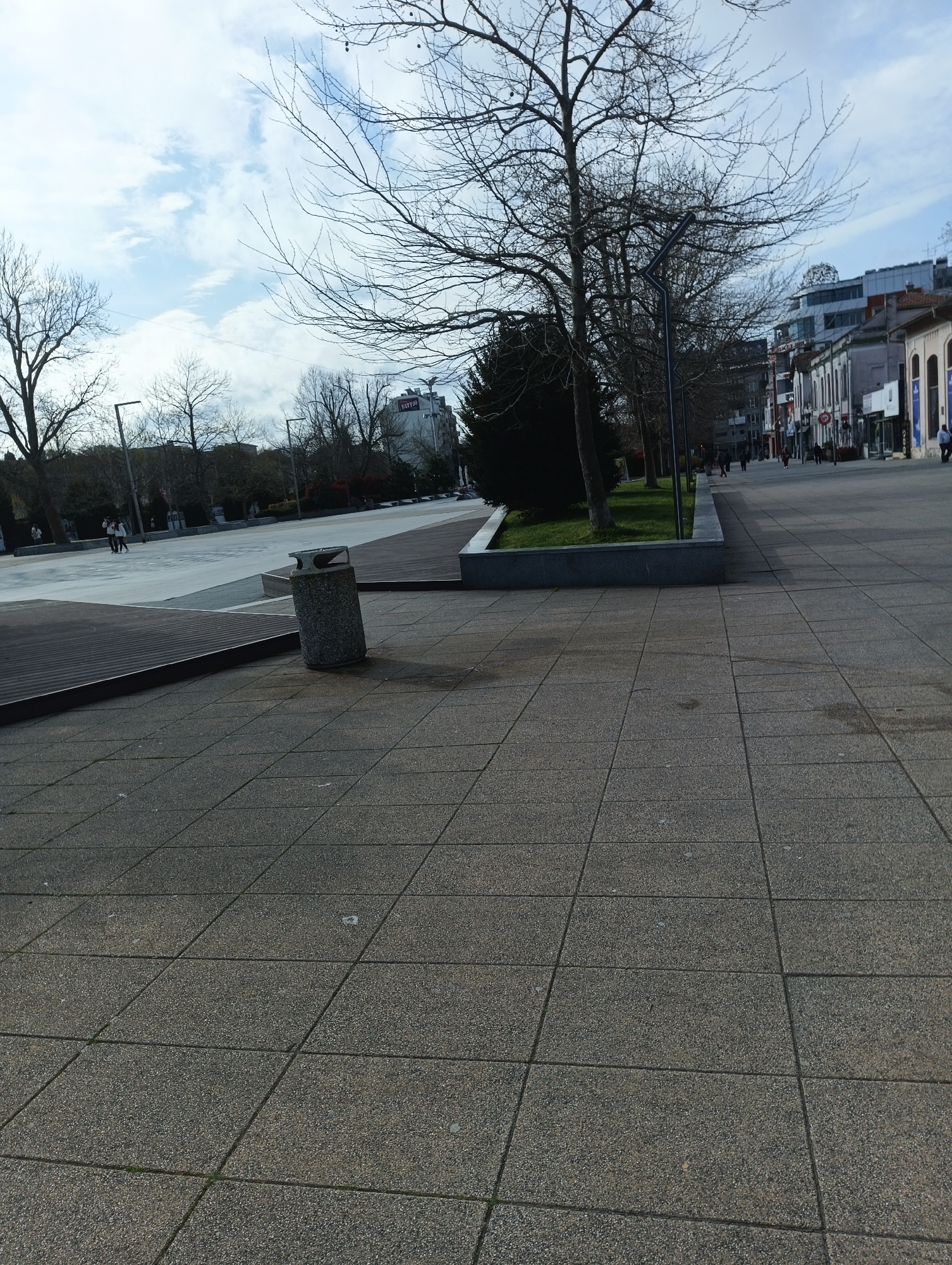 This image shows a public square in Burgas, Bulgaria. The square is paved with grey, square paving stones. A cylindrical trash receptacle is located to the left of the frame. A raised concrete planter contains trees and grass. In the background are buildings, some with commercial storefronts, and bare trees. A few people are walking in the distance. The sky is overcast with patches of blue.