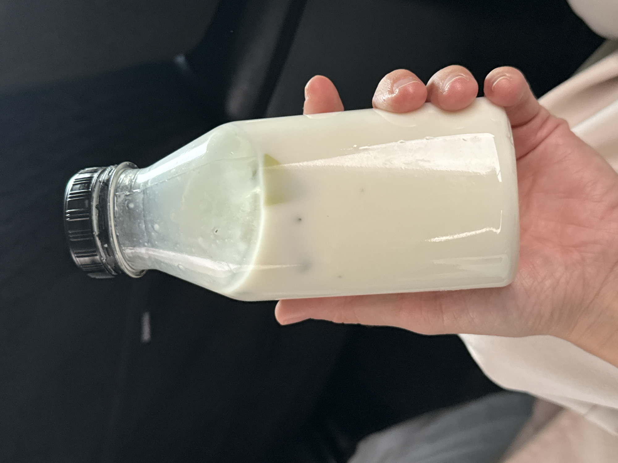 A hand holds a bottle of white liquid, possibly yogurt or milk, containing small green and black particles. The bottle has a black cap. The background shows a dark, possibly car interior, and a glimpse of a white garment. The location, Meshcheryaki, Russia, is not evident from the image.