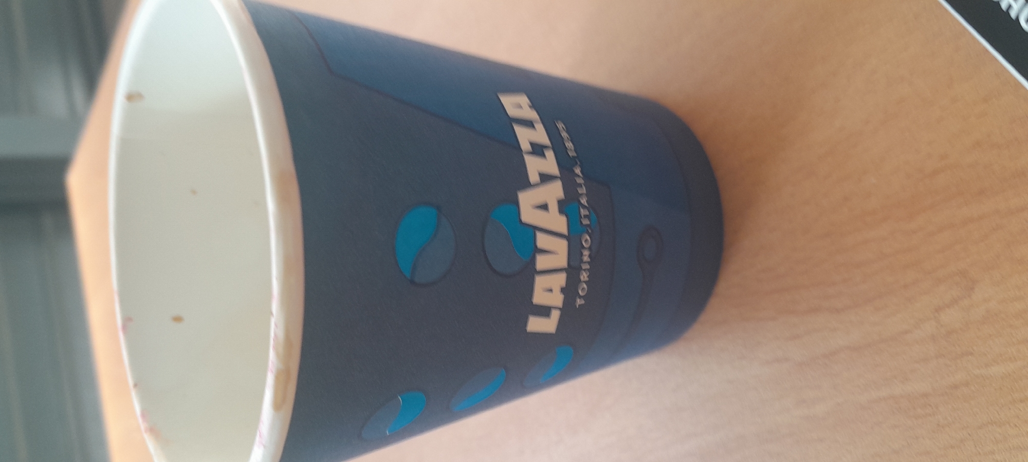 A close-up shot captures a blue paper coffee cup with the Lavazza logo and "TORINO, ITALIA, 1895" printed in white and light gray lettering. The cup is decorated with light blue oval shapes. The cup is placed on a light brown wooden surface, likely a table. The background is blurred, showing a gray surface above the table. No people or actions are visible in the image. The scene appears to be an indoor setting, possibly a cafe or office space. There is no information about the location in Sofia, Bulgaria.